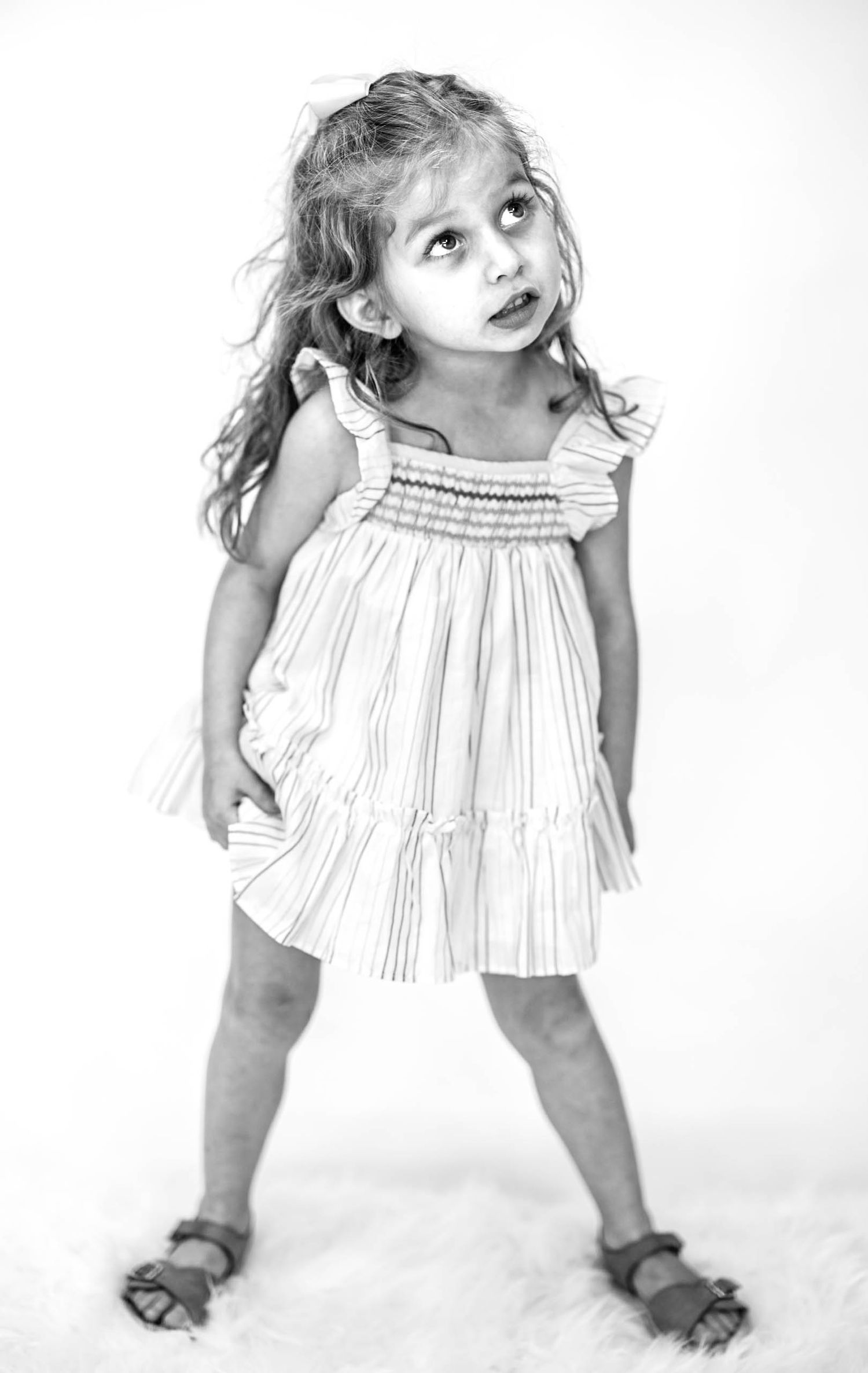 Black and white photo of a cute toddler girl in a smocked dress and sandals, looking up with curiosity.