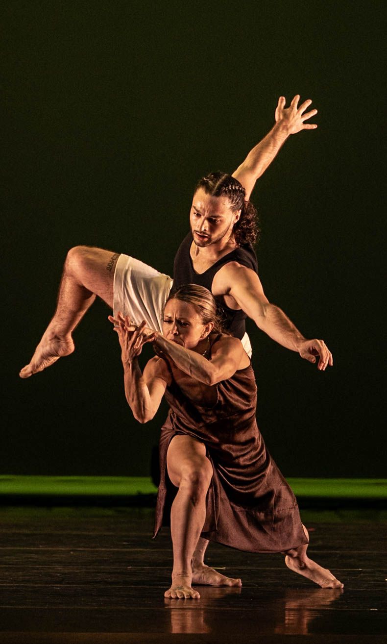 Two contemporary dancers perform a dramatic lift on a dark stage, one balanced across the other's shoulders with an arm extended.