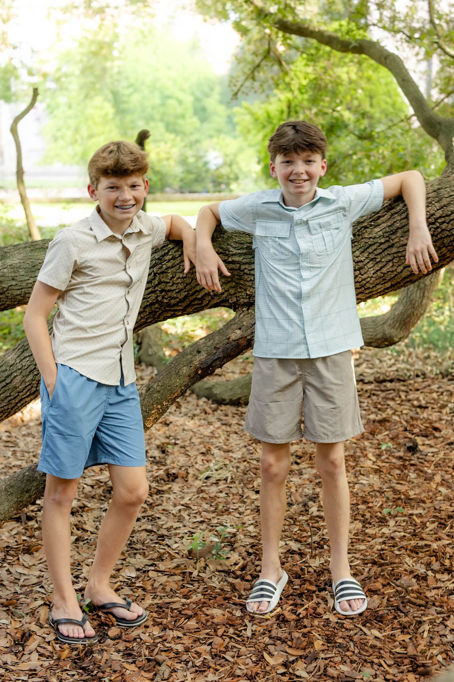 Two smiling boys lean against a fallen tree in a sunny park, wearing button-up shirts and shorts.