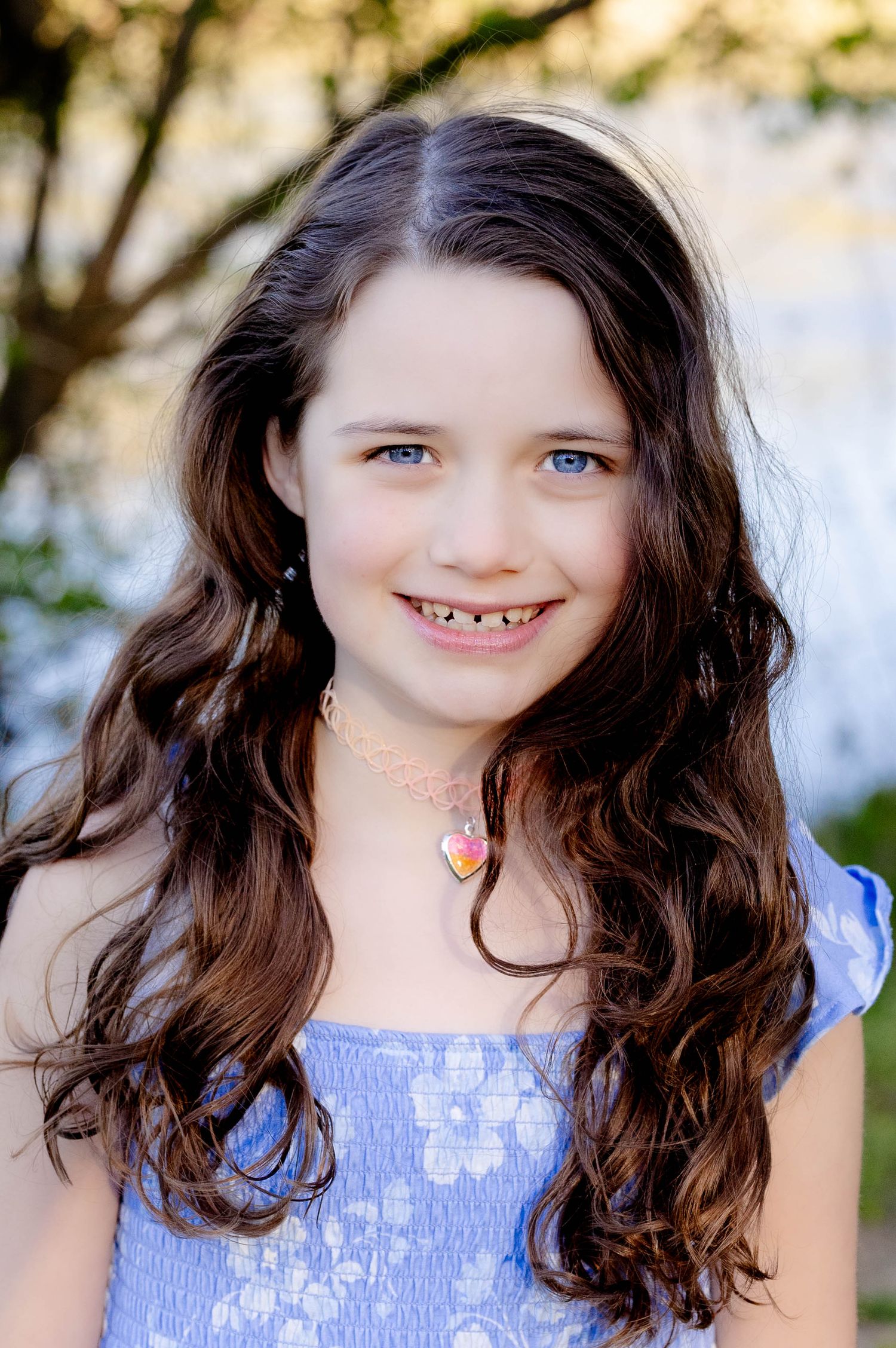 A young girl with blue eyes and curly brown hair smiles wearing a heart necklace and blue top.