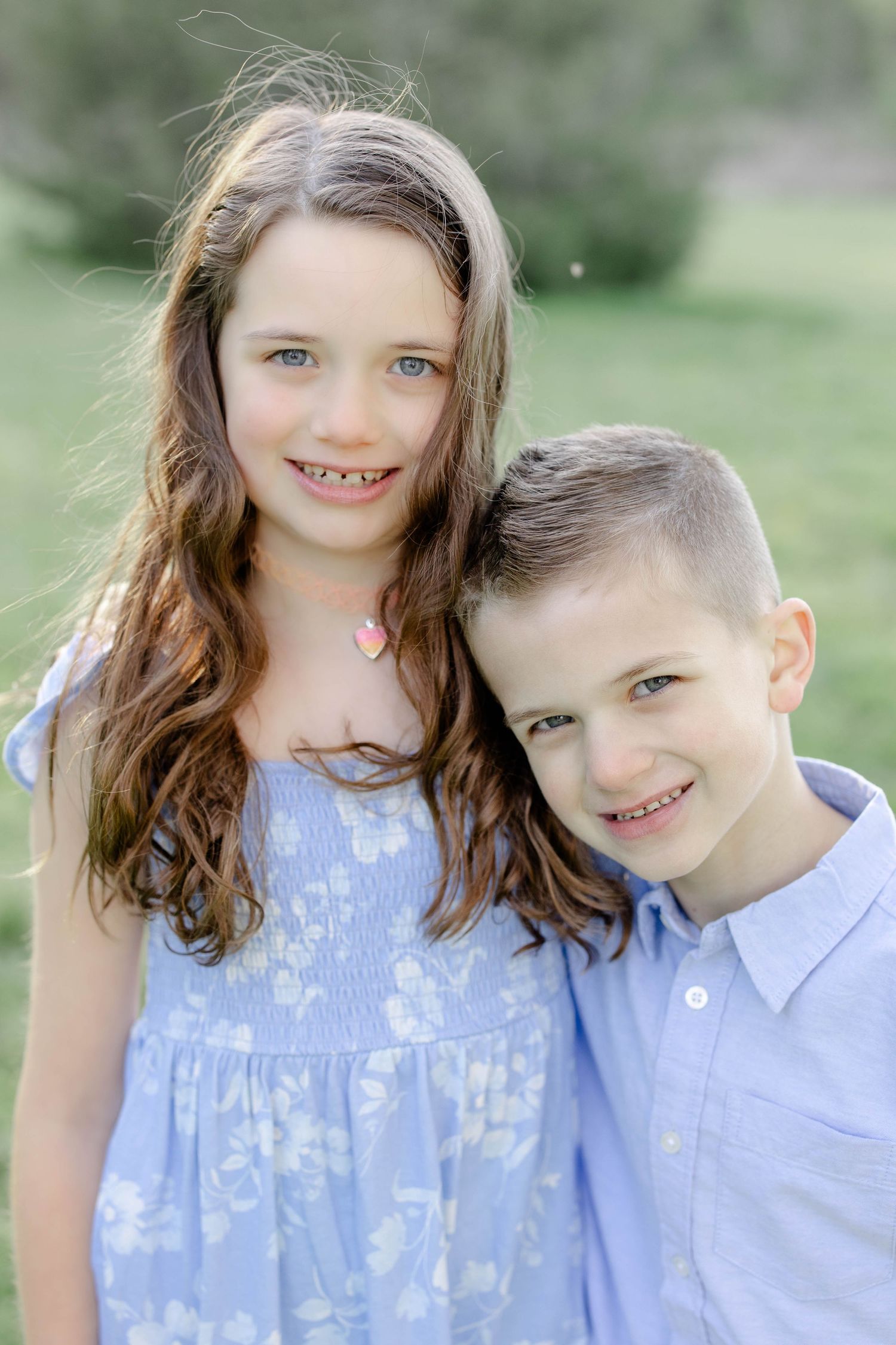 Two siblings in blue outfits smiling together outdoors in a green field.