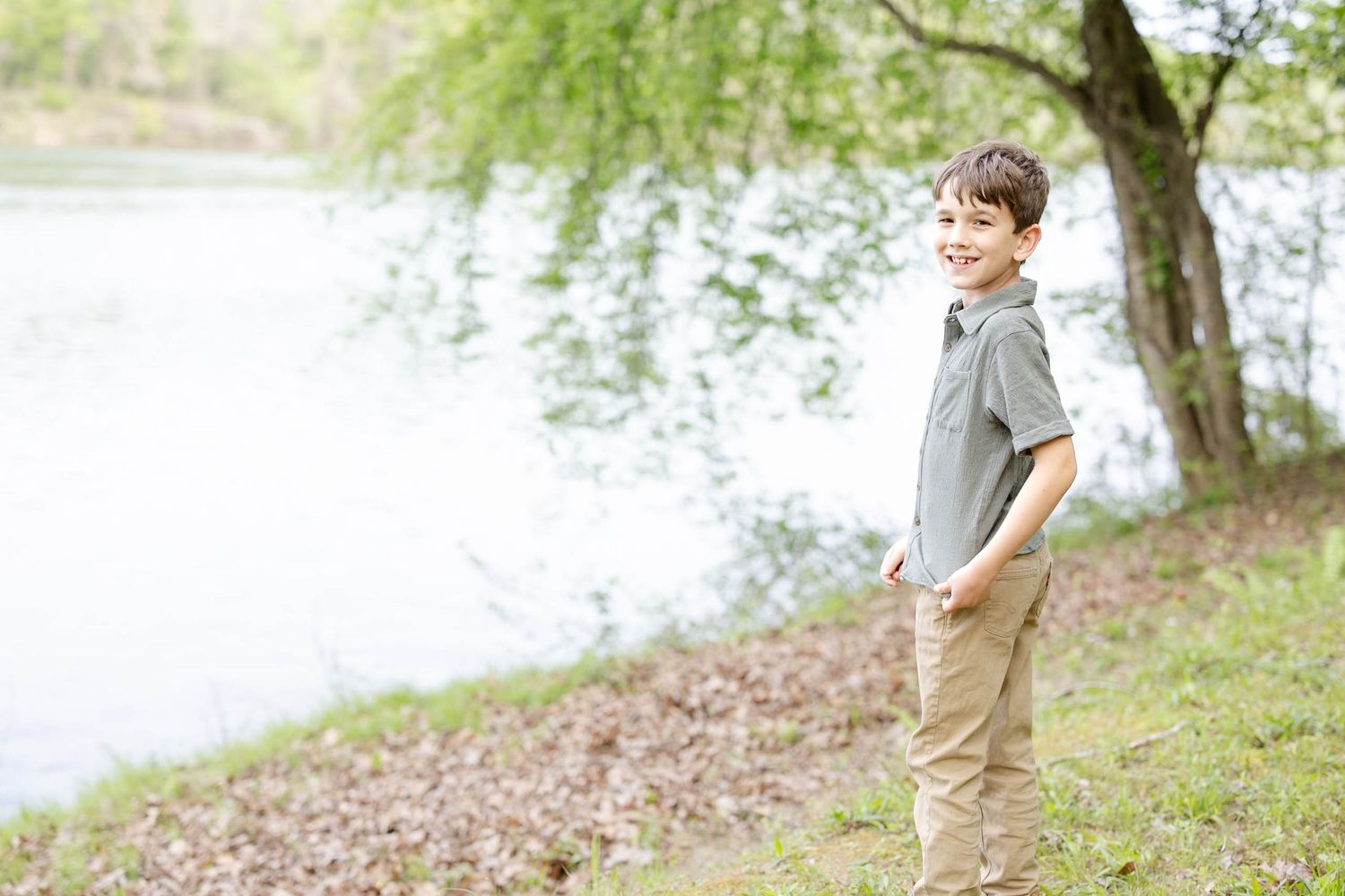 Smiling young boy standing near a lake under a willow tree in a lush green park.