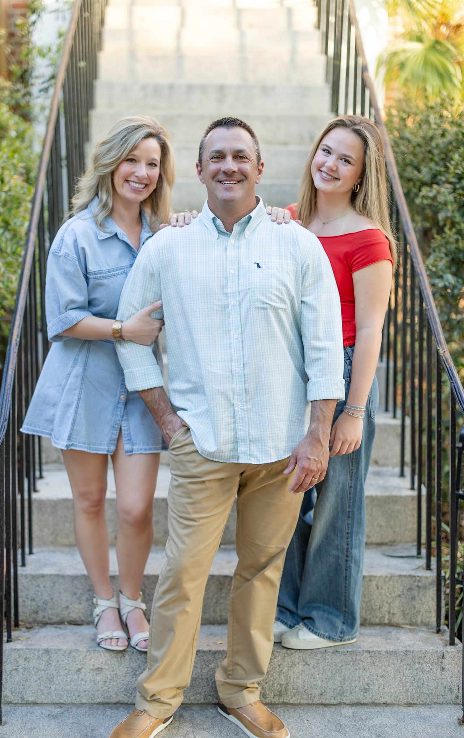 Family of three smiling on outdoor stairs, mother in denim dress, father in white shirt, teen in red top.