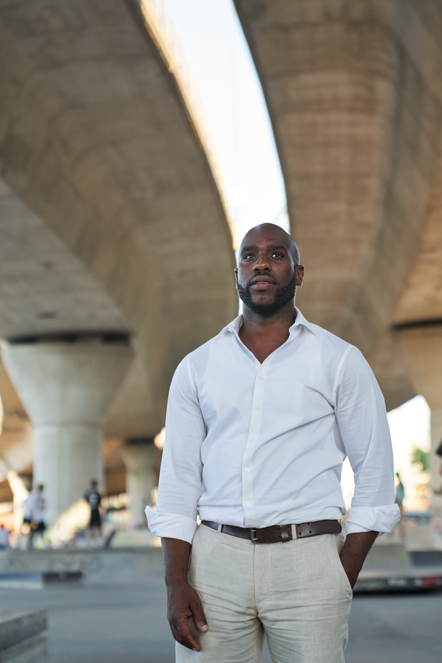 A confident Black man in a white shirt and beige pants stands under a concrete bridge overpass in an urban setting.