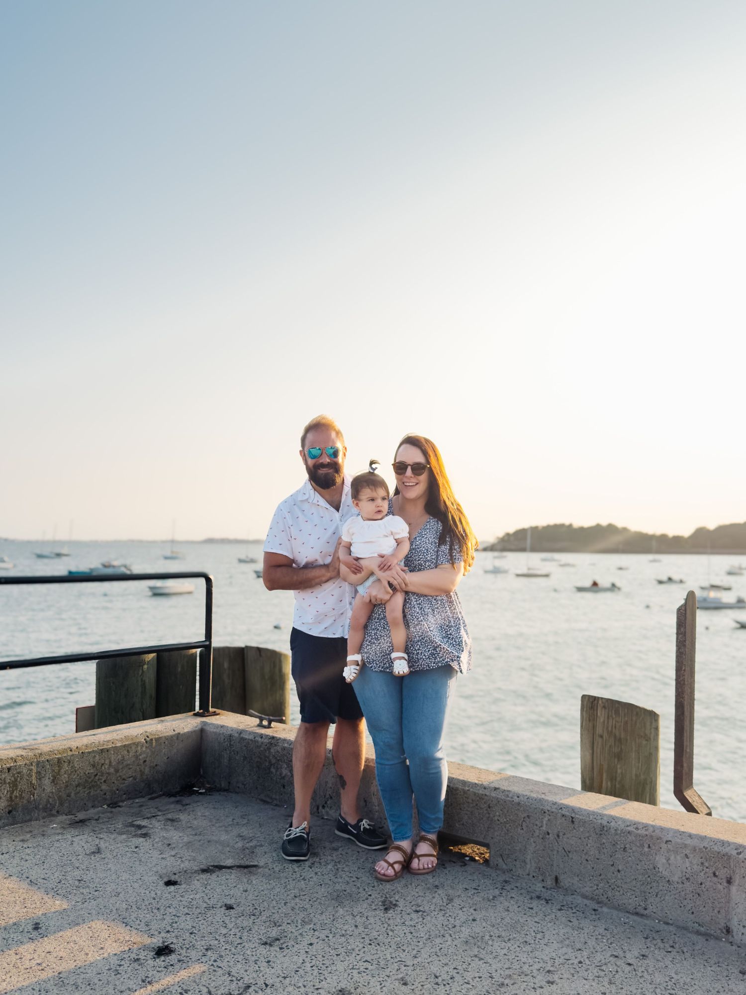 Family of three with baby posing on waterfront pier at golden sunset with boats behind.