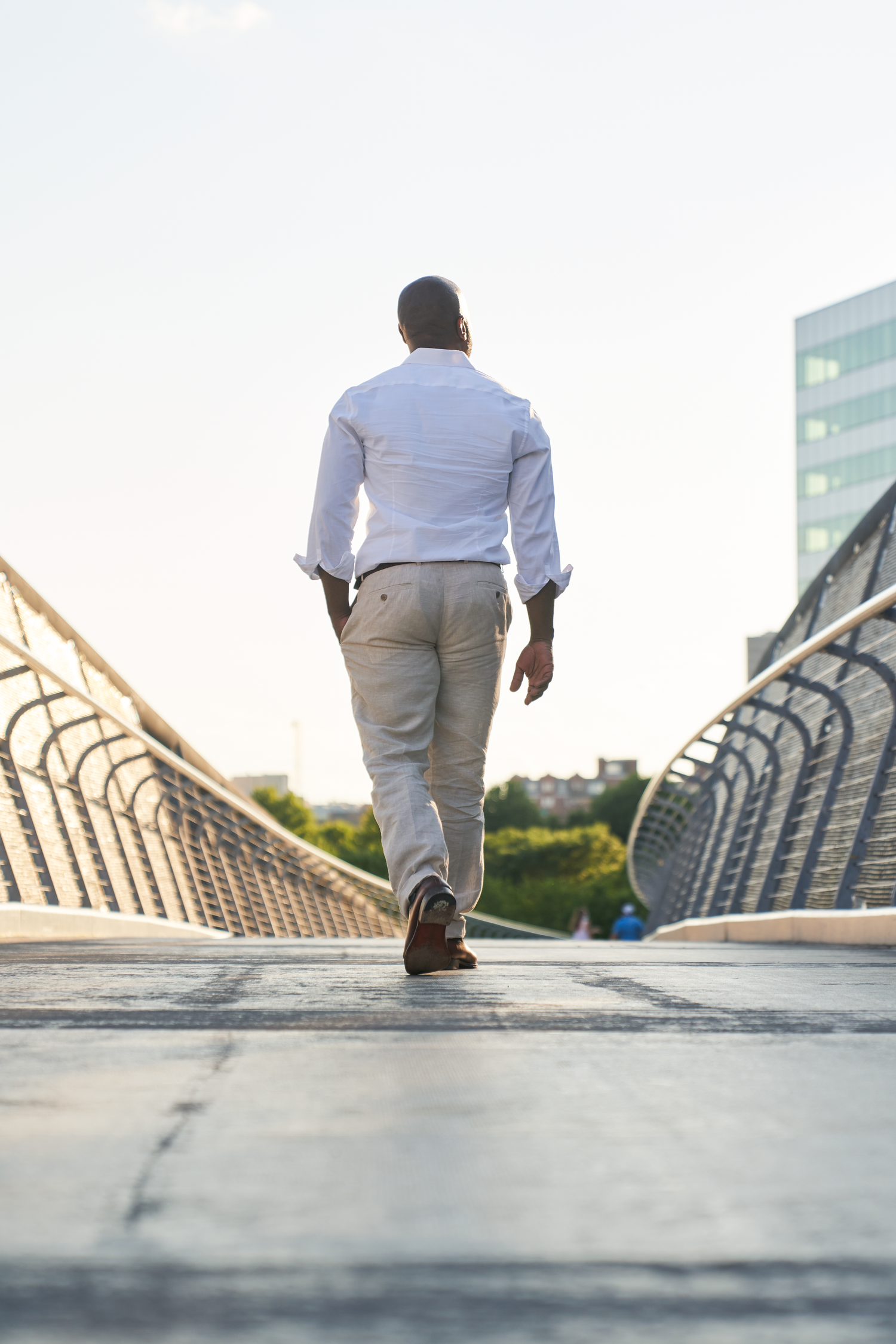 Black man in white shirt and gray pants walking away on modern bridge in urban environment.
