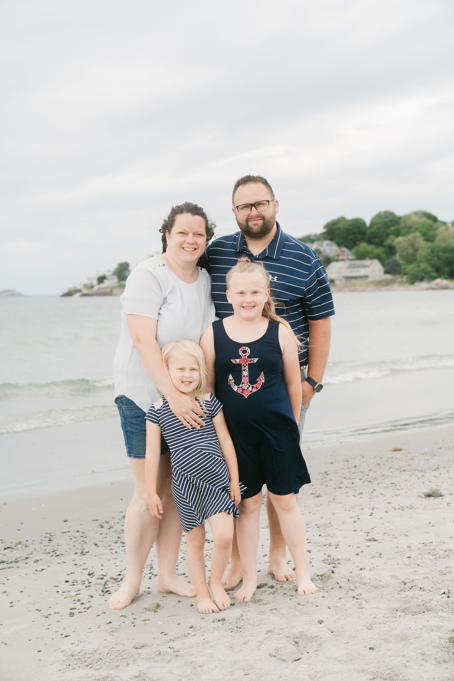 Family of four posing barefoot on a sandy beach, two adults and two girls in nautical outfits, with ocean waves in the background.