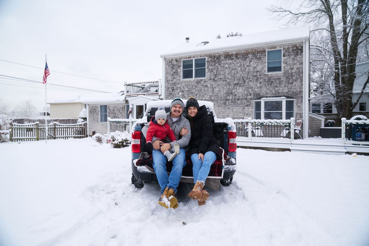 Happy family with a dog sitting on a snowy pickup truck bed in front of a winter home.