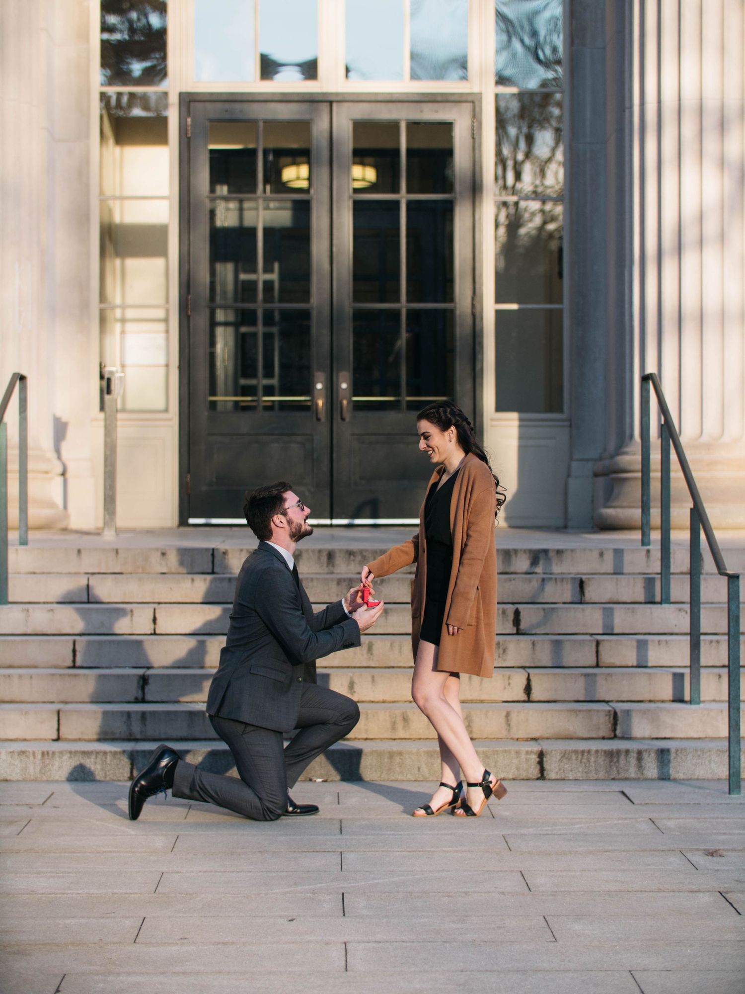 Man proposing on one knee to woman on stone steps outside a classical building.