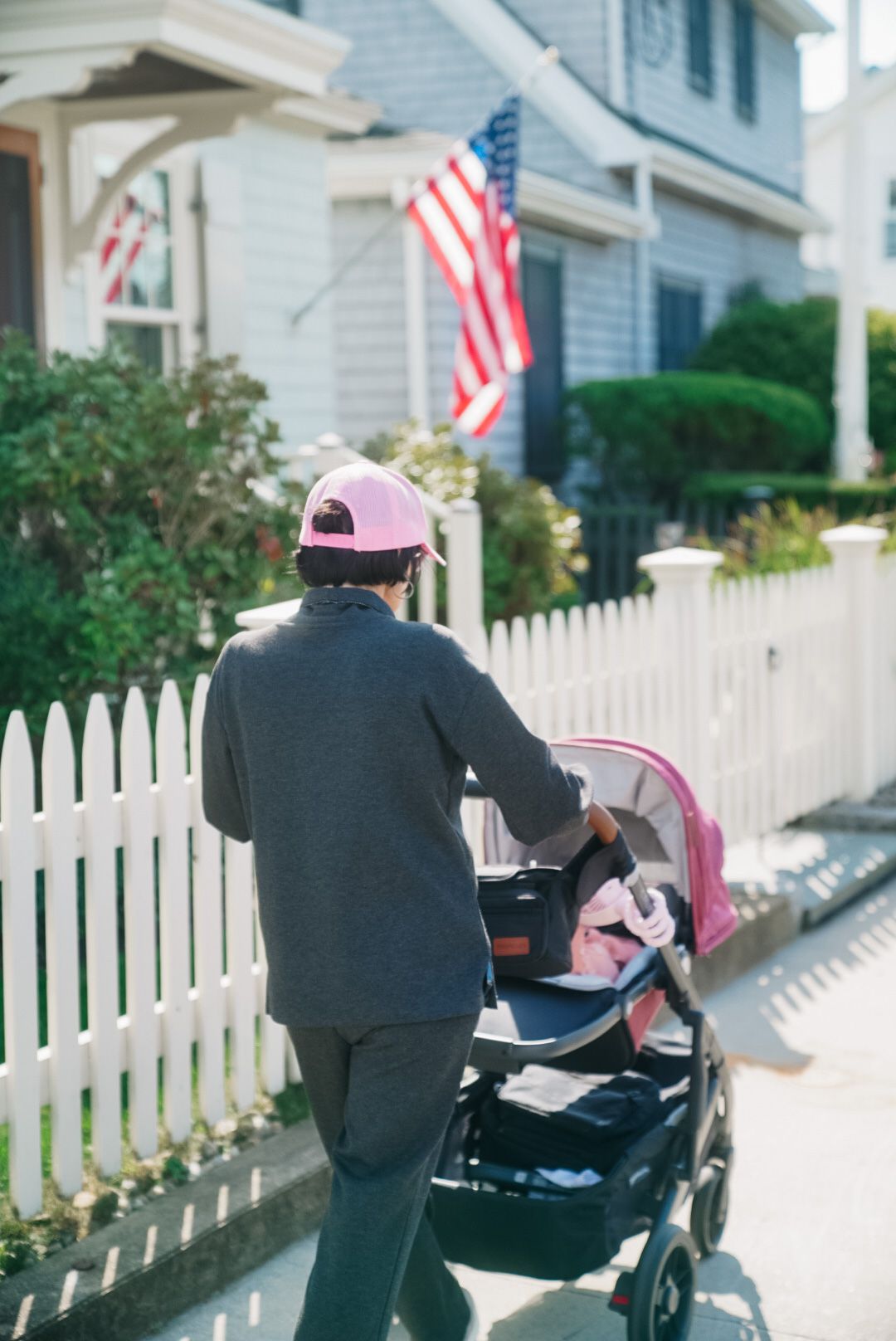 A person in a pink cap pushes a stroller past a white picket fence with an American flag.
