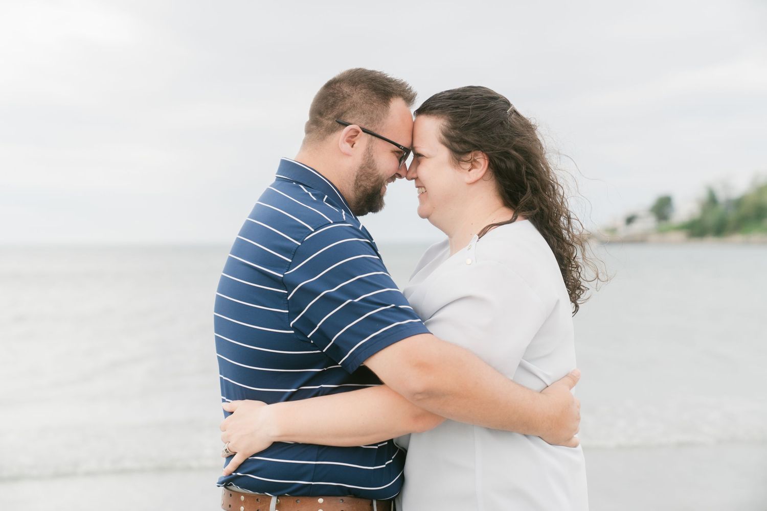 A couple embraces and touches foreheads romantically on a beach with calm water in the background.