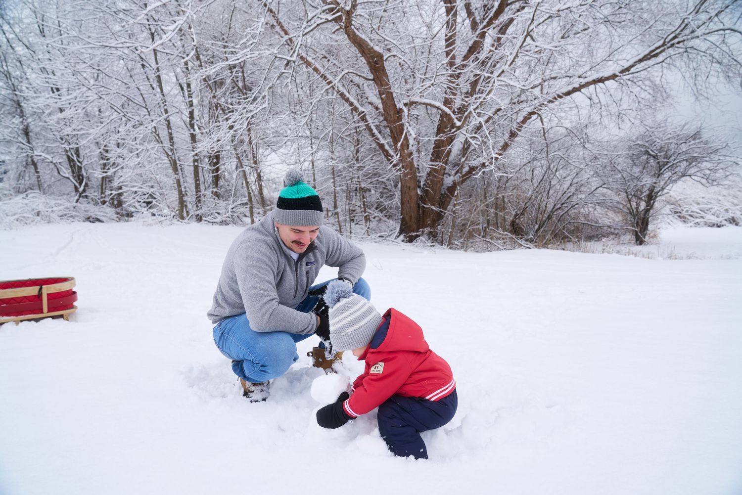 Father and young child in red snowsuit playing together in fresh snow near frost-covered trees.