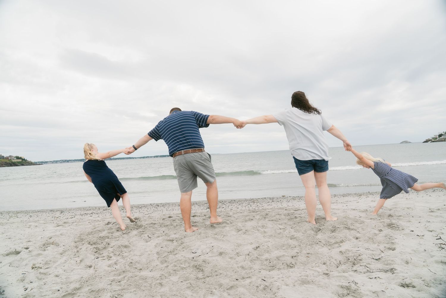 Family of four holding hands on a sandy beach, facing the ocean on a cloudy day.