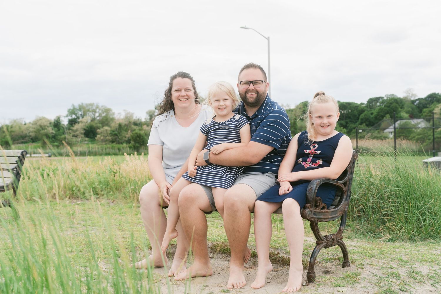 Family of four sitting on a bench near sandy dunes and tall grass on a cloudy day at the beach.