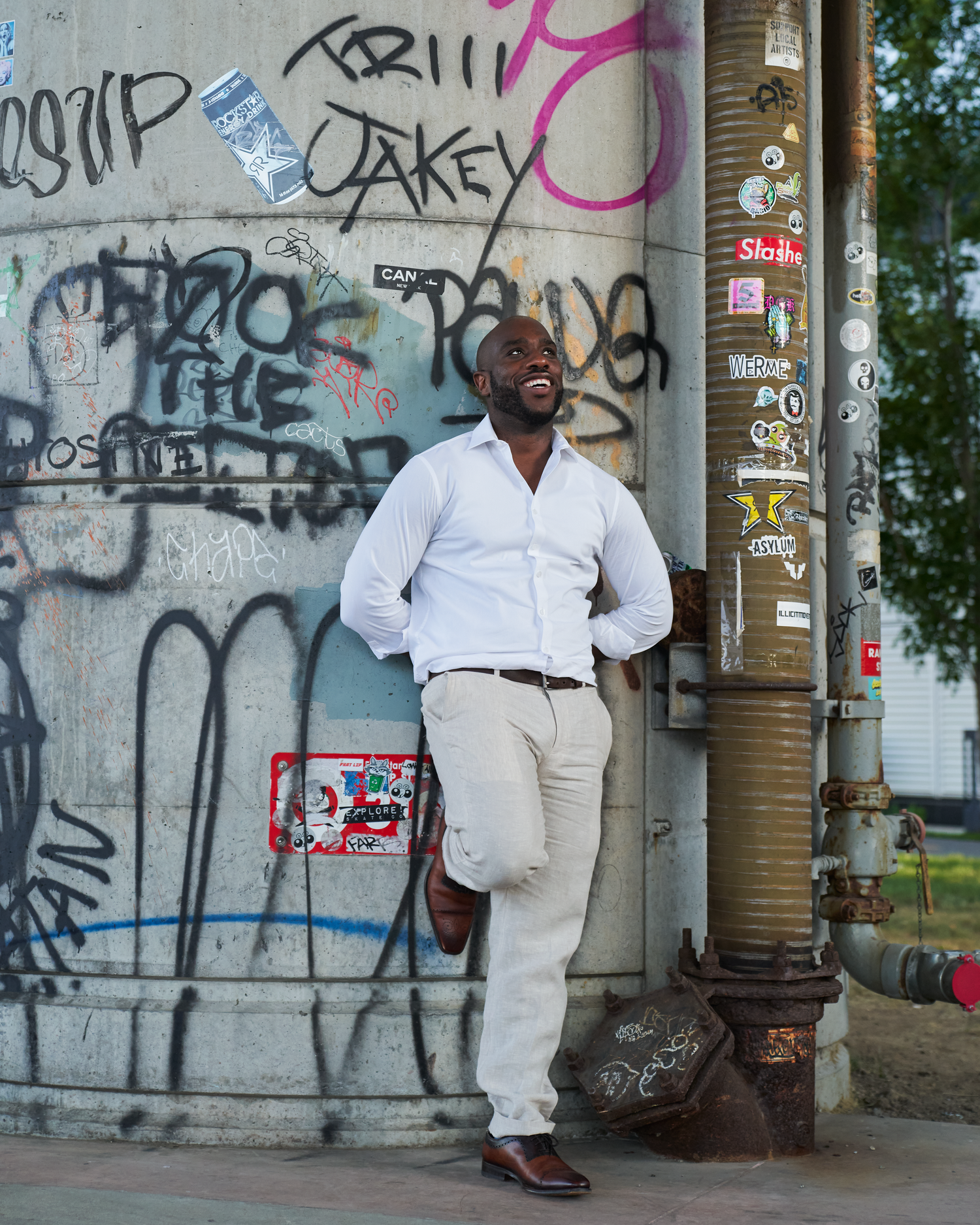 A smiling man in white shirt and khaki pants leans against a graffiti-covered wall near an urban pole.