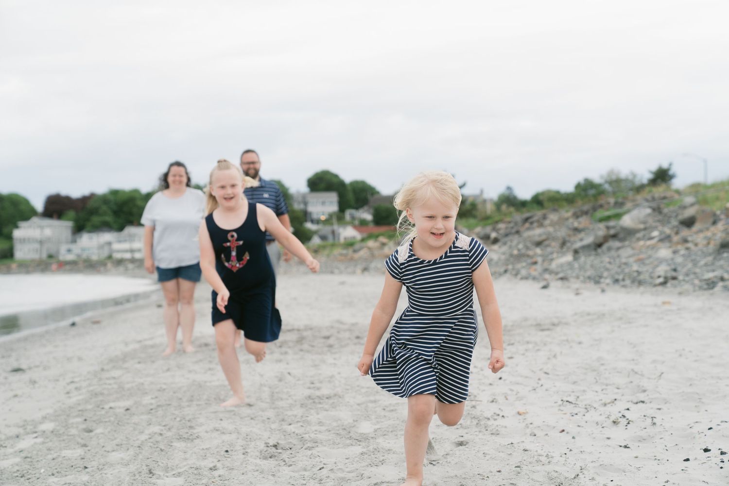 Two girls running on a sandy beach while parents walk behind on a cloudy day.