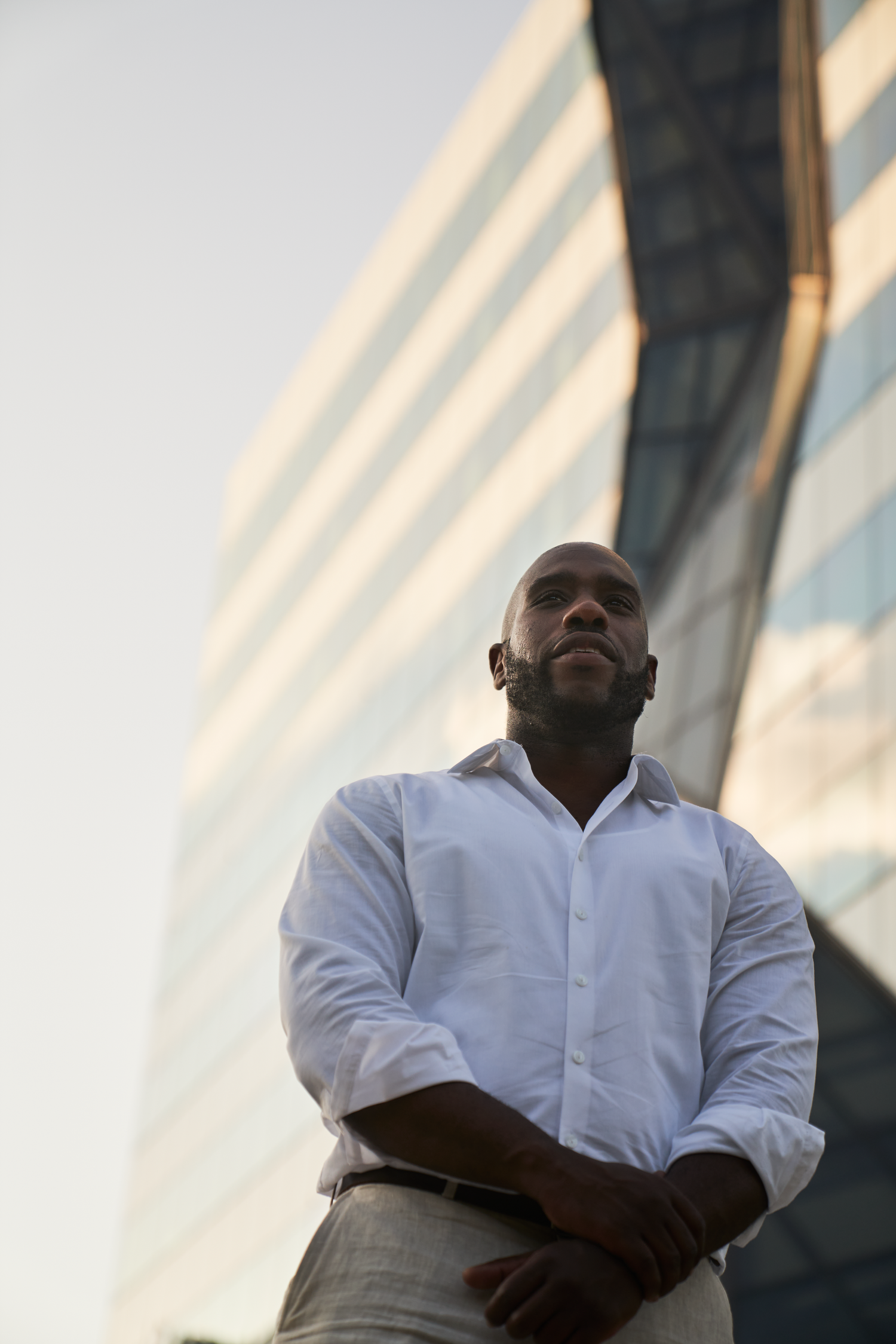 Confident Black businessman in white shirt standing outdoors near modern glass building, looking up thoughtfully.