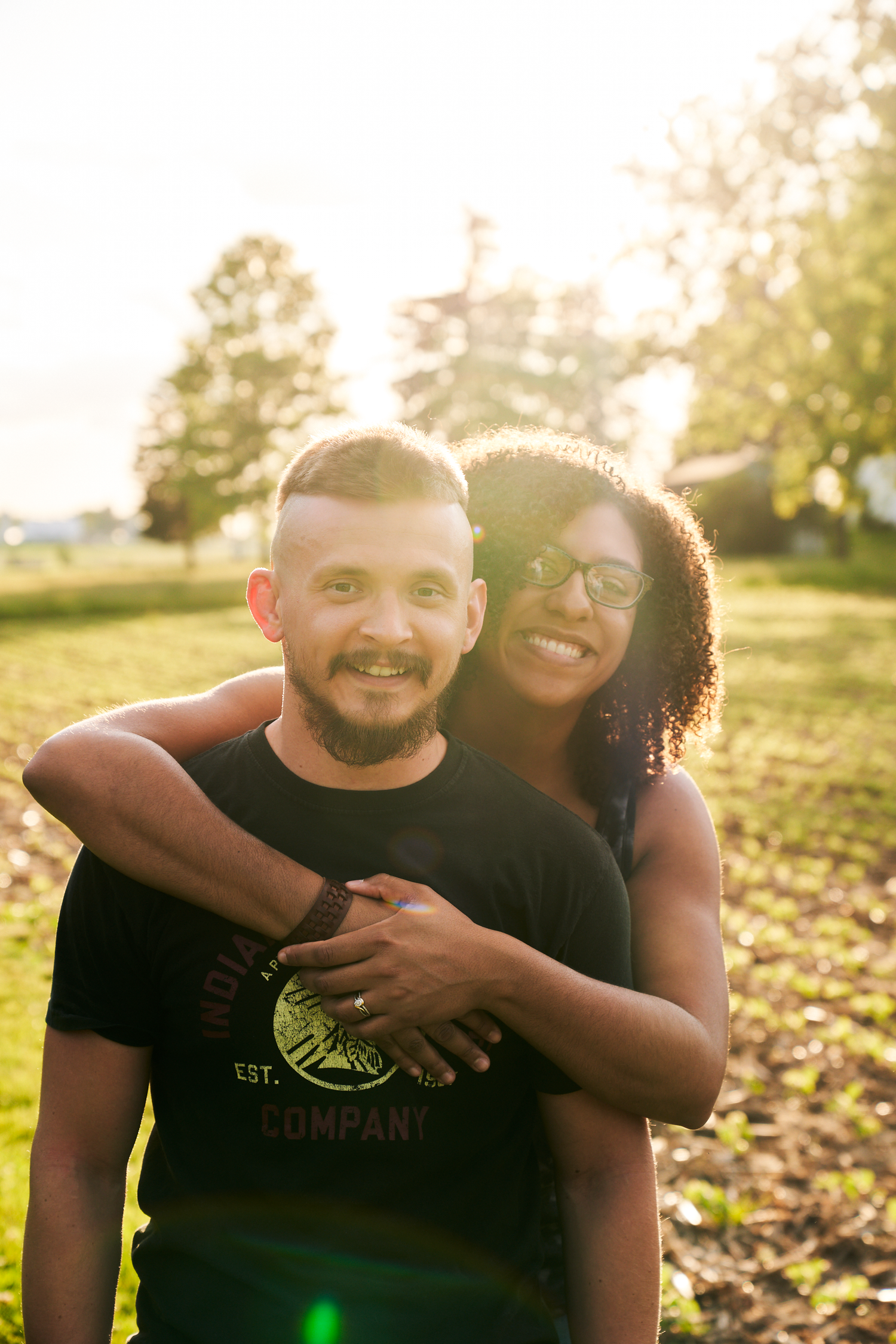 Smiling couple embracing in a sunlit green field, woman hugging man from behind.