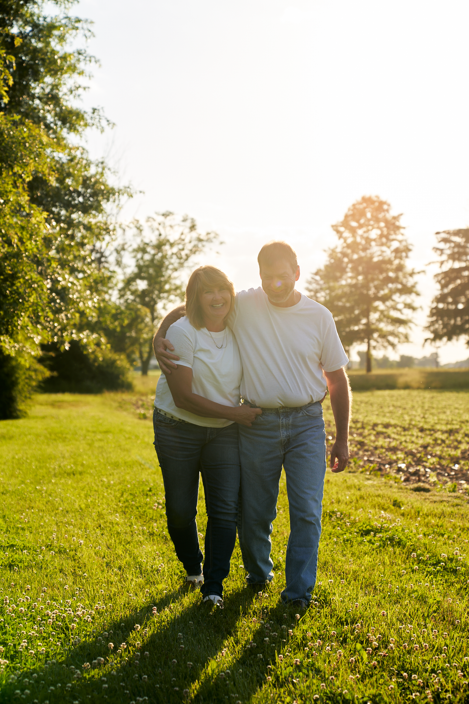 Couple walking arm in arm through a sunny green field at golden hour.