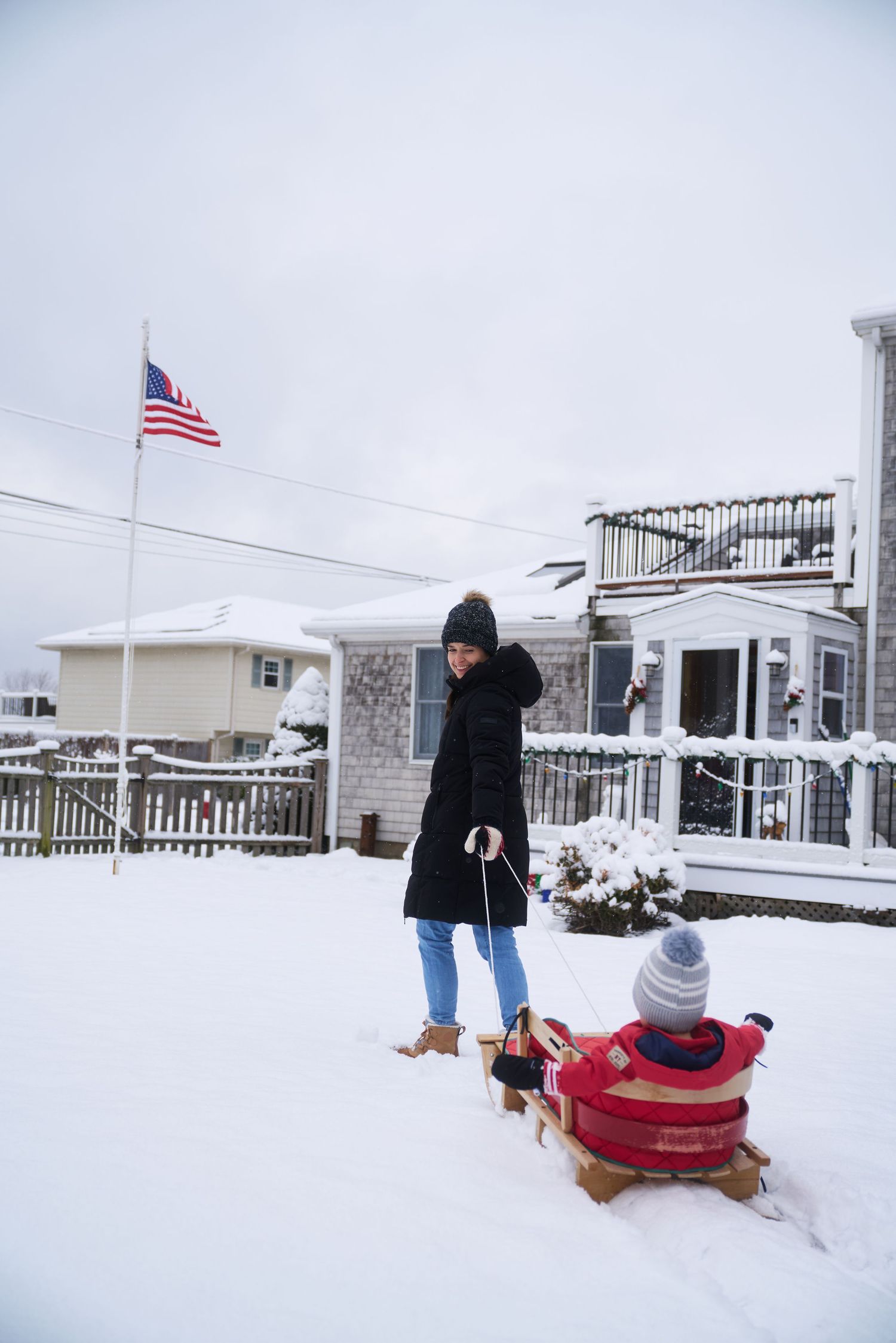 Woman in black jacket pulling child on red sled in snowy yard with American flag.
