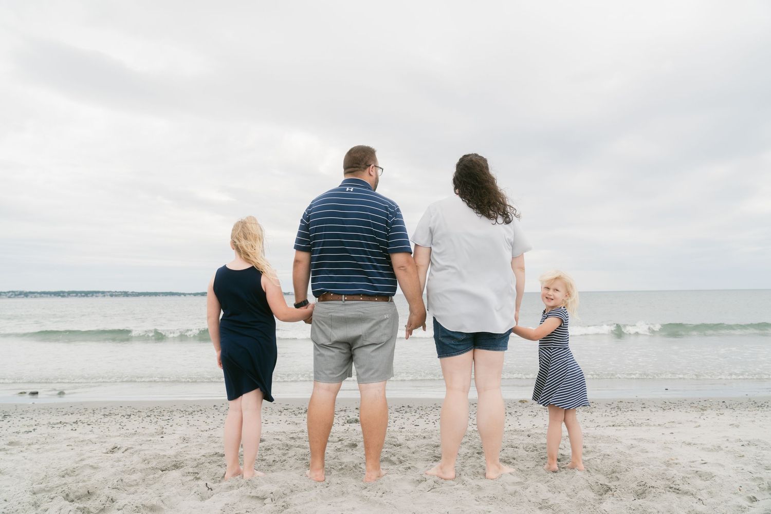 Family of four holding hands facing the ocean on a sandy beach.