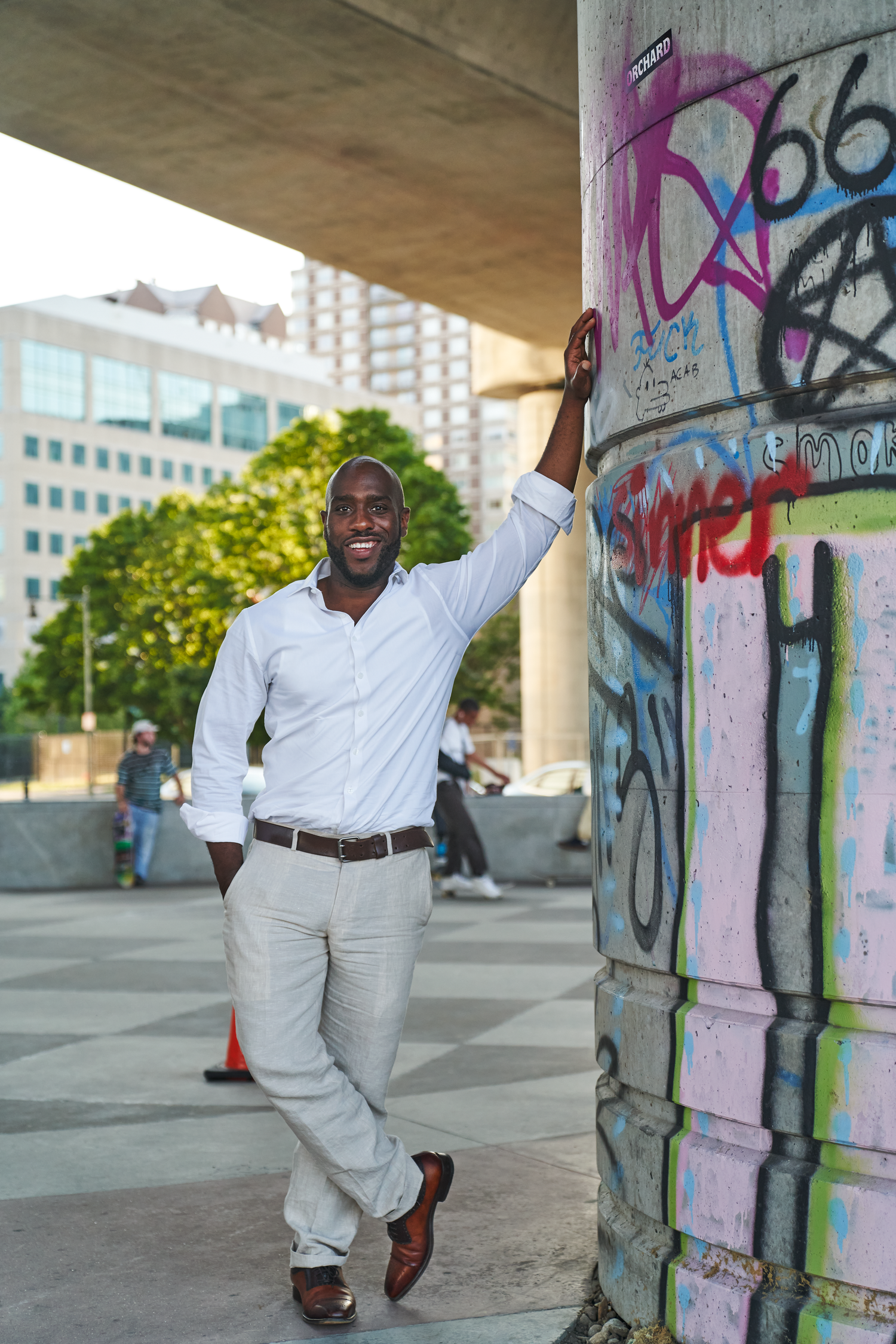 Smiling Black man in white shirt and gray pants leaning against graffiti-covered pillar in urban setting.
