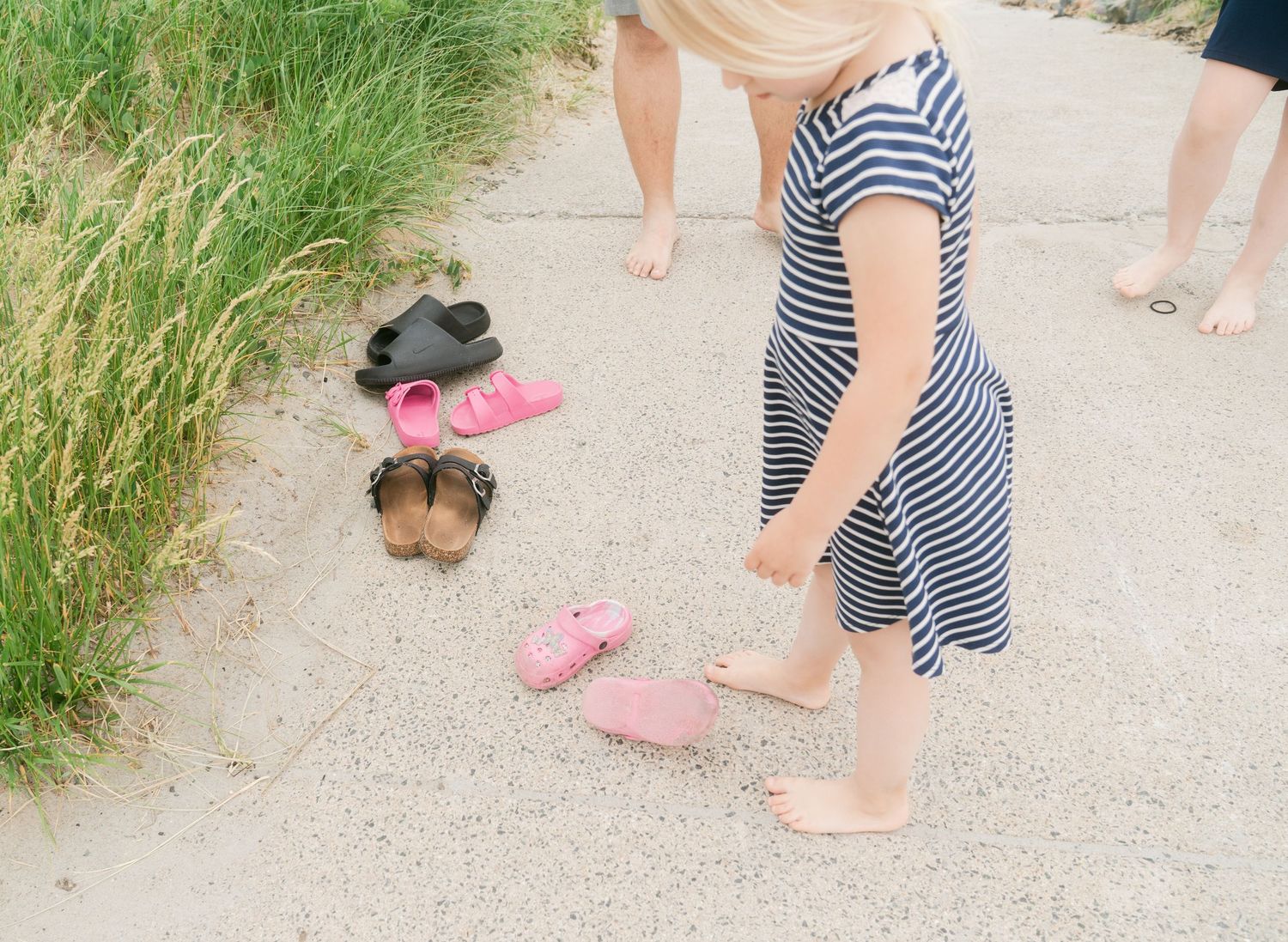 Girl in striped dress putting on pink sandals on sandy path with multiple pairs of shoes nearby.