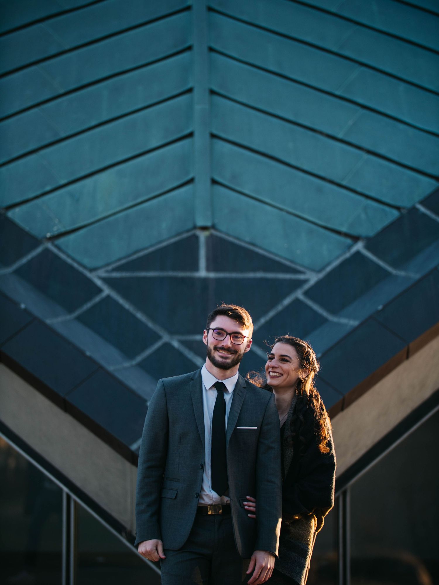 Stylish couple posing together in formal attire in front of geometric architectural structure.