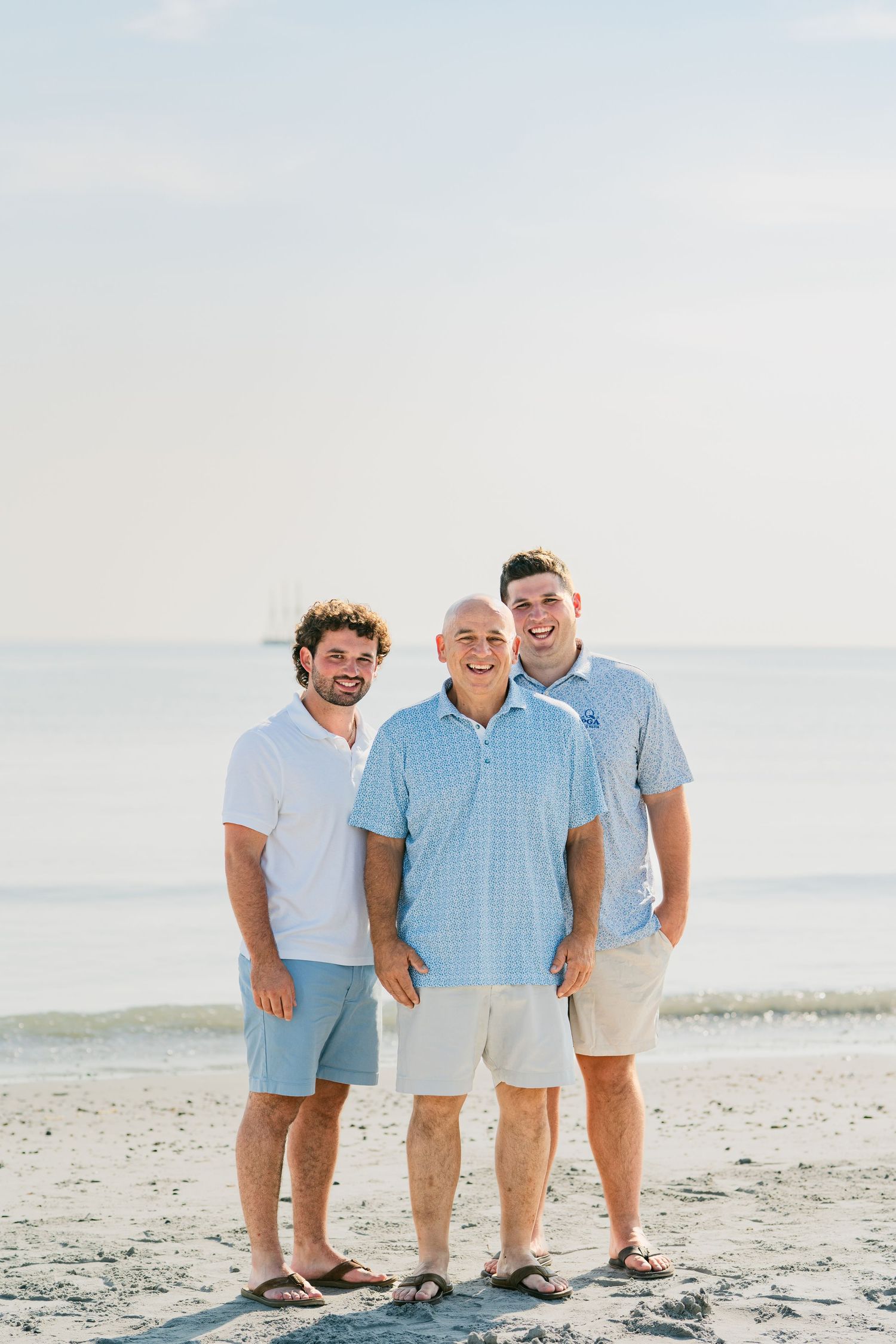 Three smiling men in casual beach attire pose together on a sandy shore with calm ocean waters behind them.