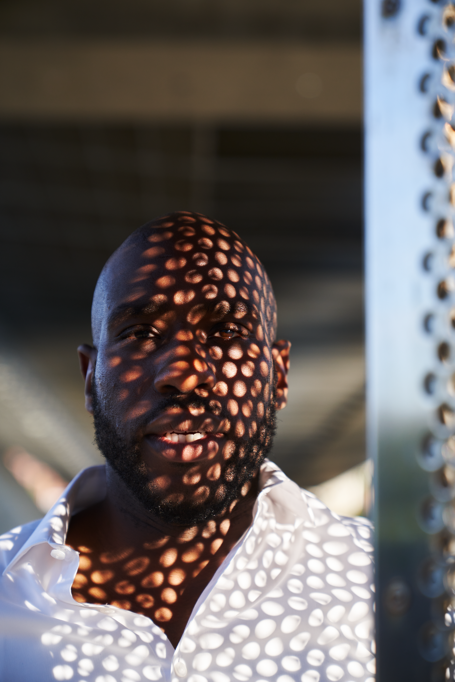 A bald Black man in a white shirt with circular light patterns casting polka dot shadows across his face outdoors.