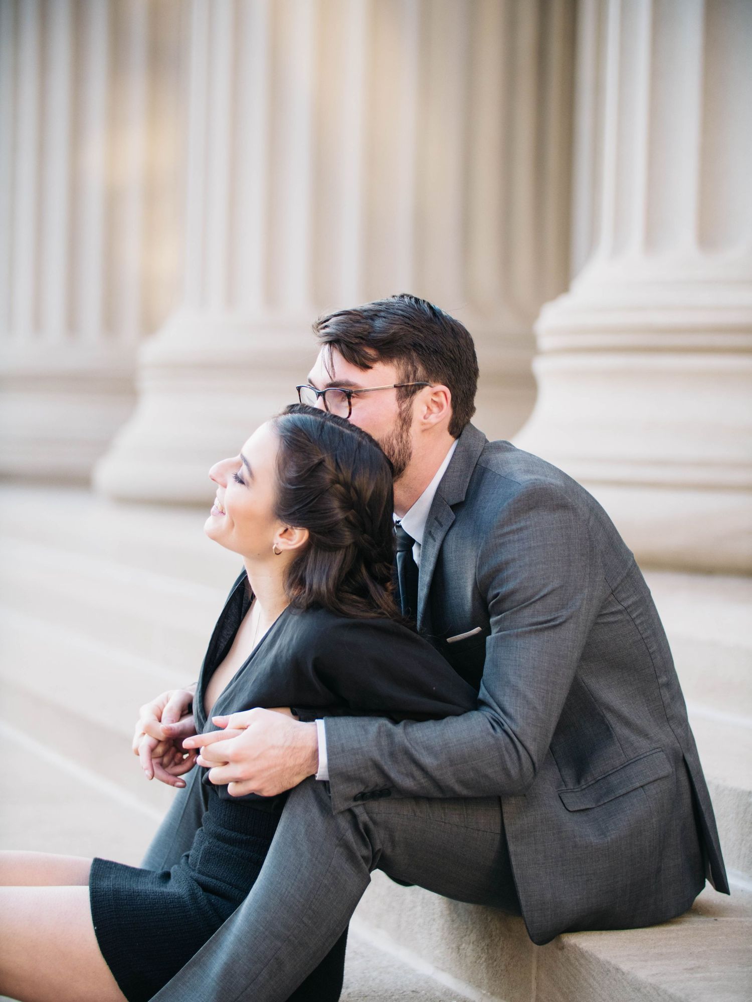 Man kissing woman on cheek while embracing on steps with large stone columns behind them.
