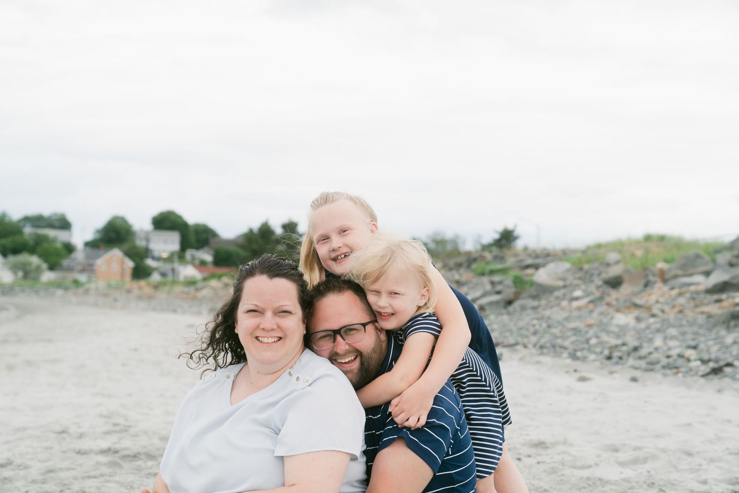 Happy family of four smiling together on a rocky beach, parents with two young blonde children on a cloudy day.