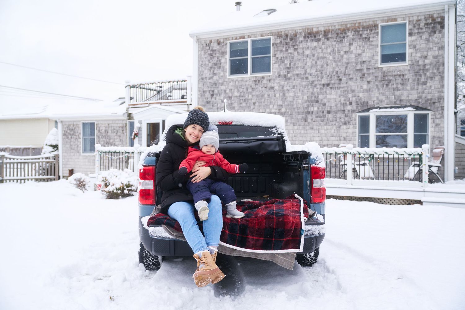Mother in black coat holding baby in red snowsuit on truck tailgate in winter snow.