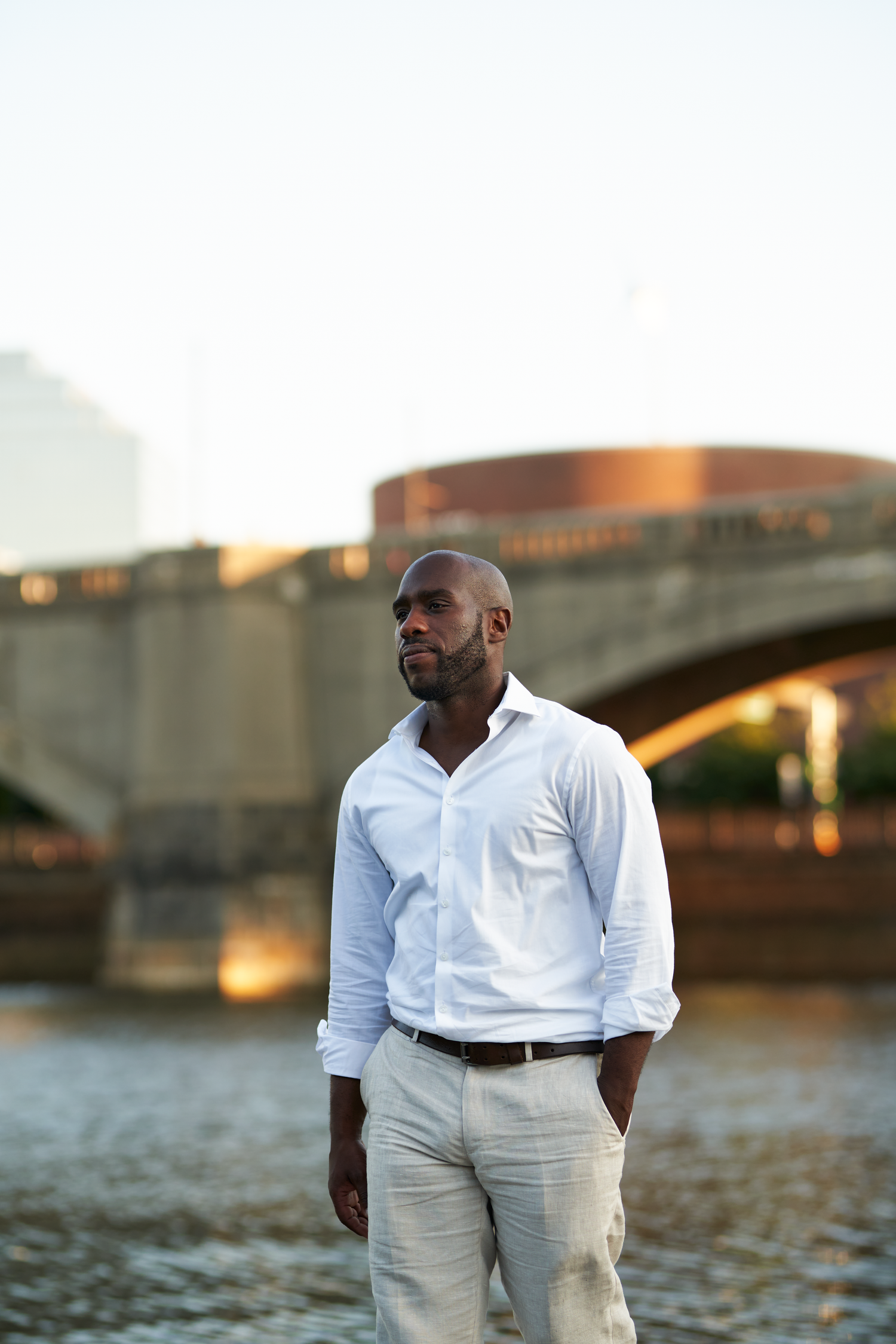 A well-dressed Black man in white shirt and khaki pants stands near a river with a stone bridge backdrop.