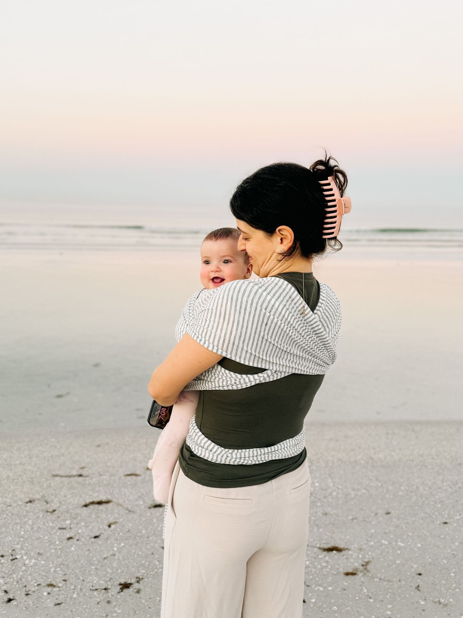 A mother holds her baby on a beach at dusk, both facing the calm ocean waves.
