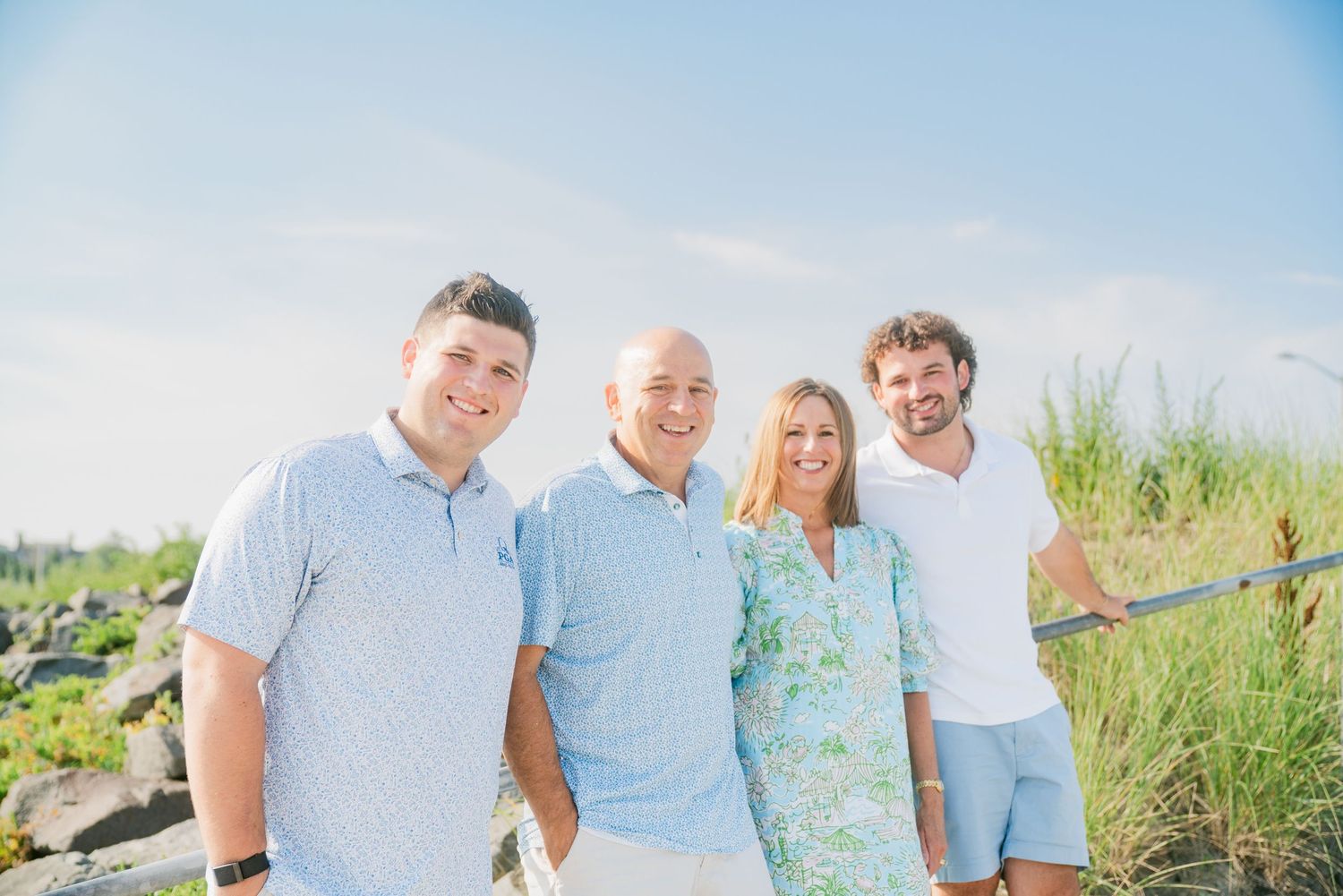 Family of four smiling together outdoors near coastal grasses on a sunny day, wearing casual summer attire.