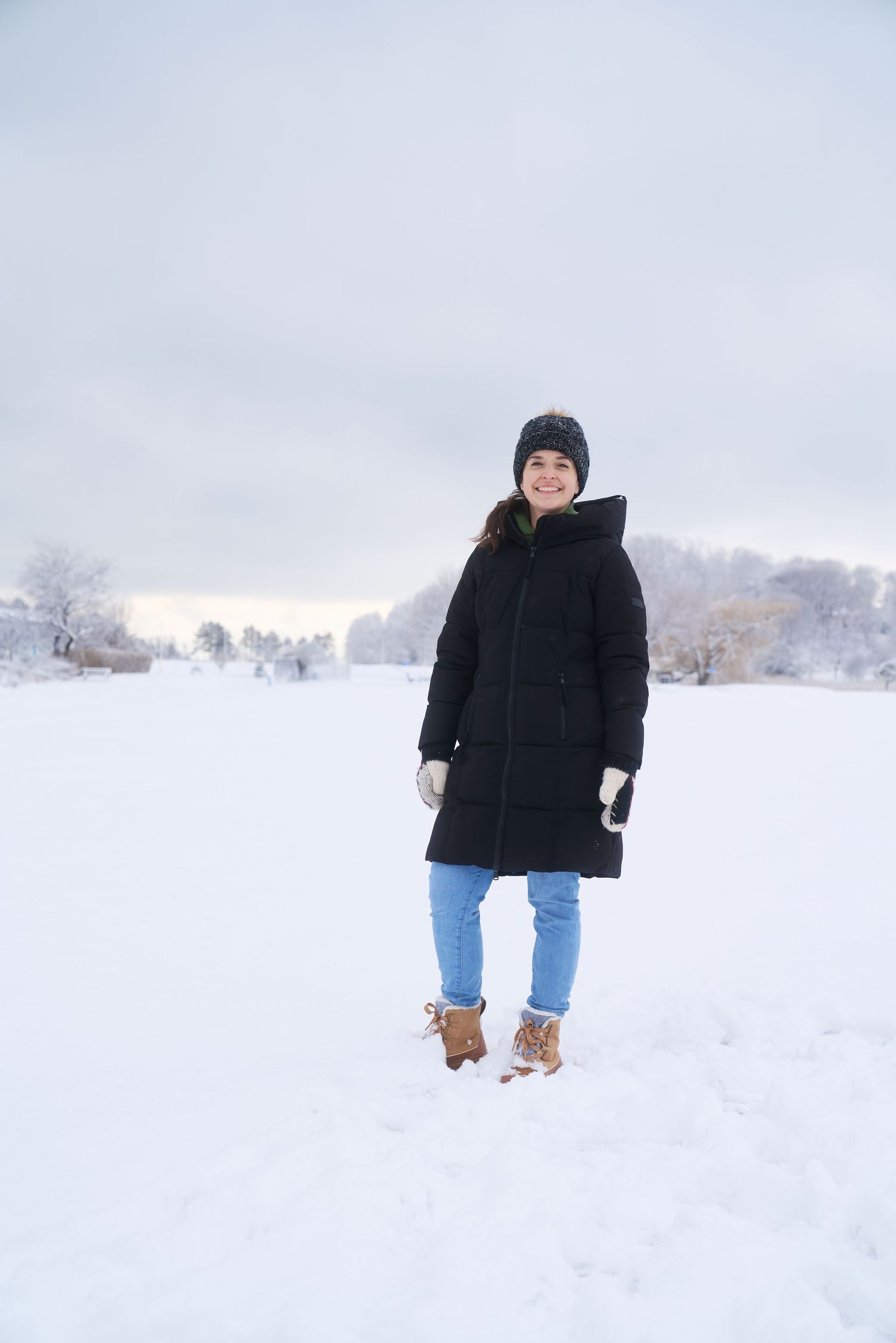 Smiling woman in black puffer coat and jeans standing in open snowy winter landscape.