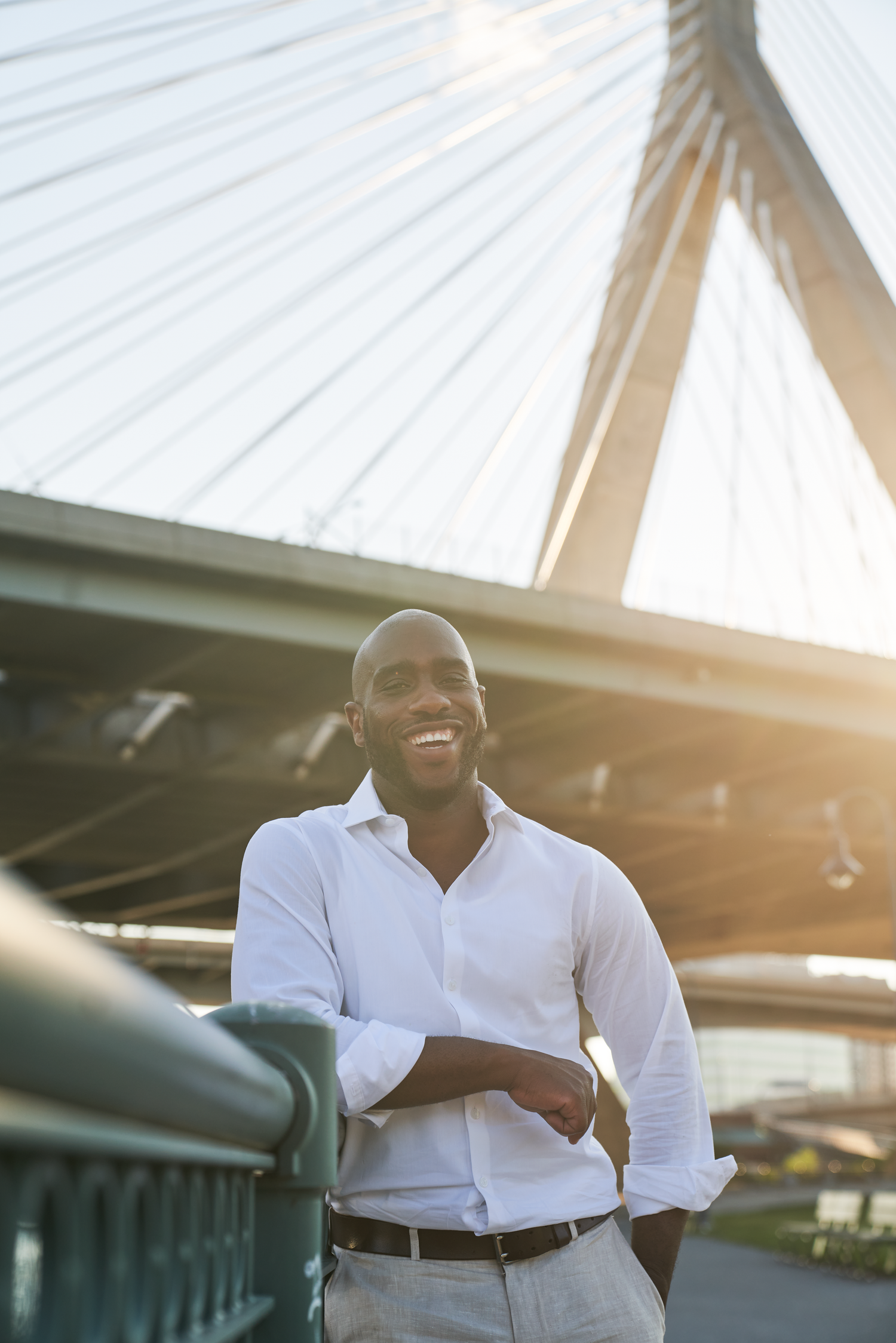 Smiling bald Black man in white shirt leaning on railing with cable-stayed bridge in sunny background.