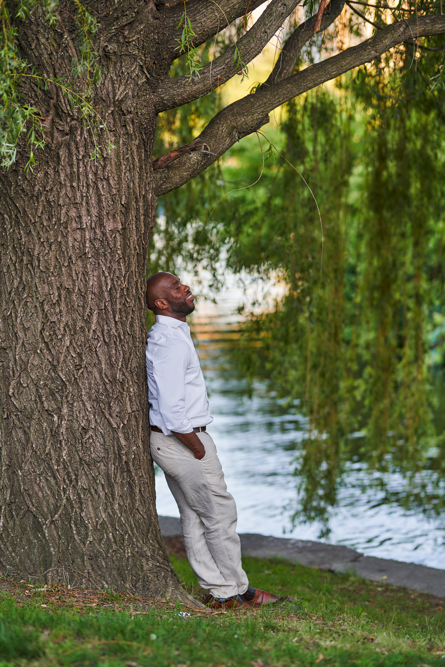 A man in a white shirt and khaki pants leans against a large tree by a serene pond in a lush park.
