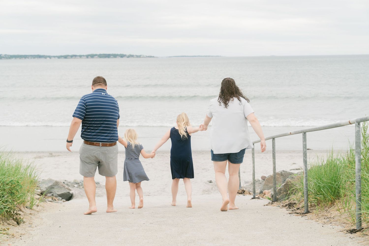 Family of four walking hand-in-hand toward a calm beach on a cloudy day, viewed from behind.