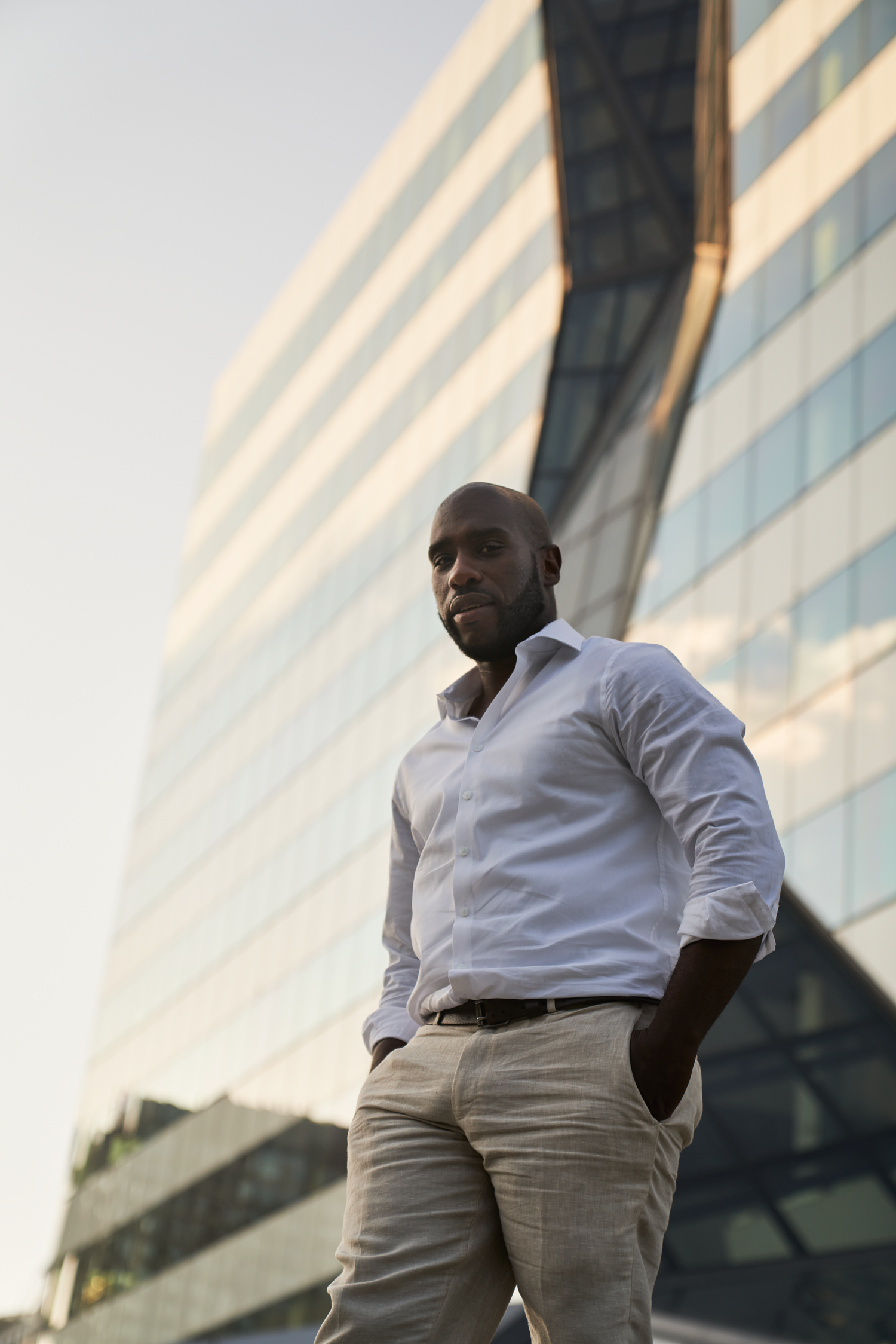 Confident Black businessman in white shirt and khakis standing outdoors in front of modern glass office building.