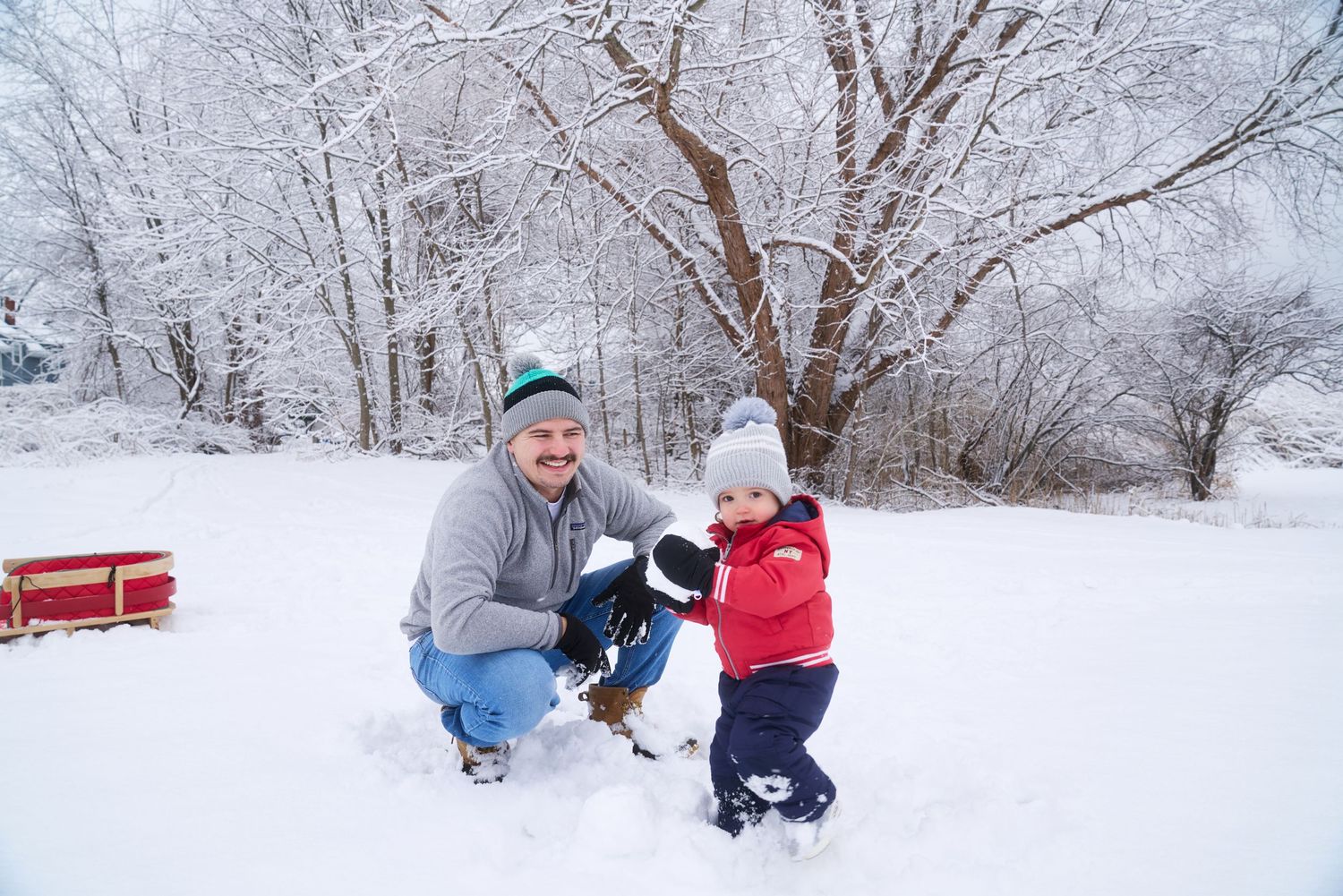 Father crouching in snow with toddler in red snowsuit near red sled in winter park.