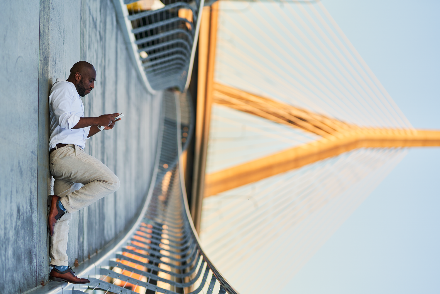 Man in white shirt leaning against bridge pillar using phone, with cable-stayed bridge in background.