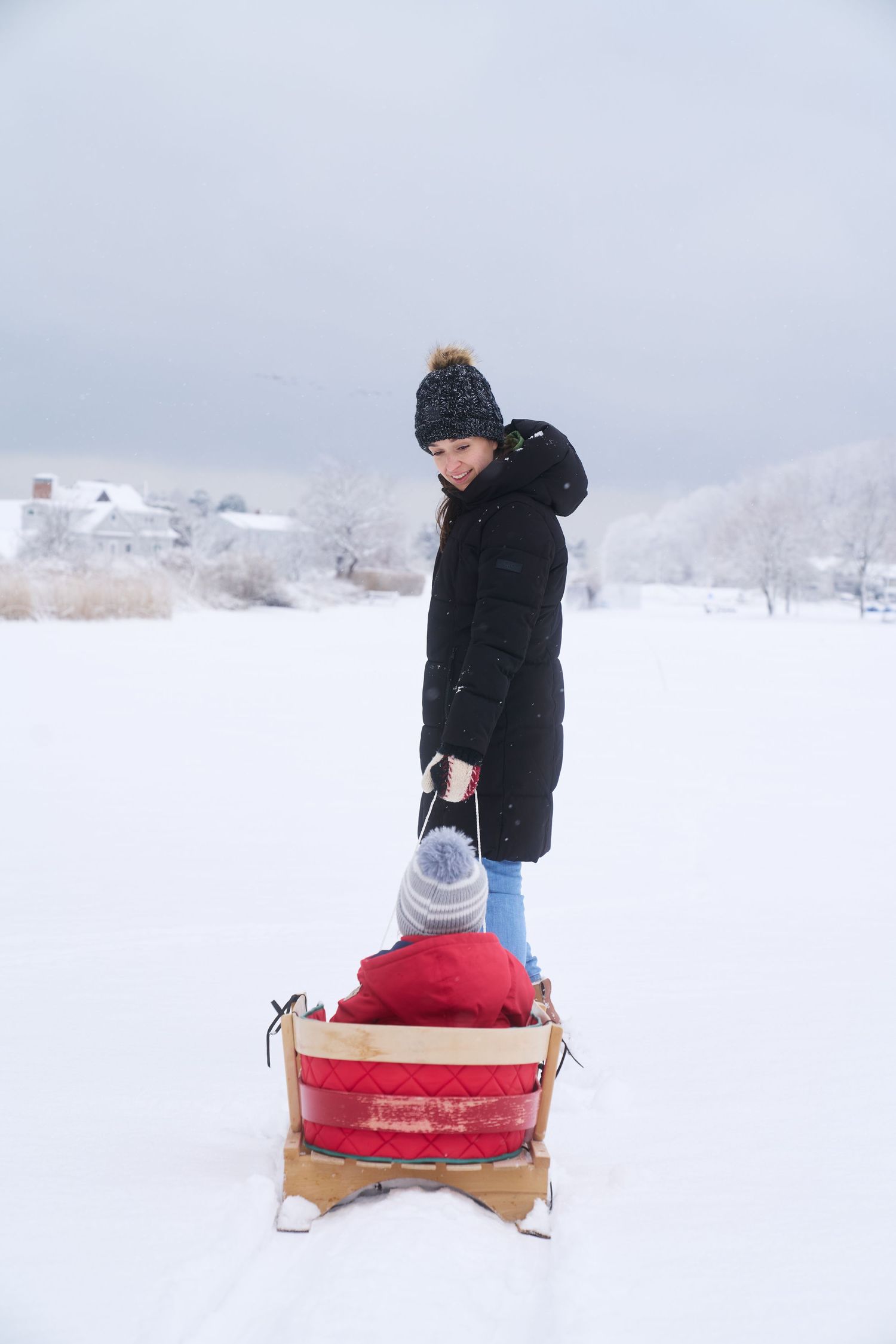 Mother in black puffer coat pulling young child on red wooden sled in snowy landscape.