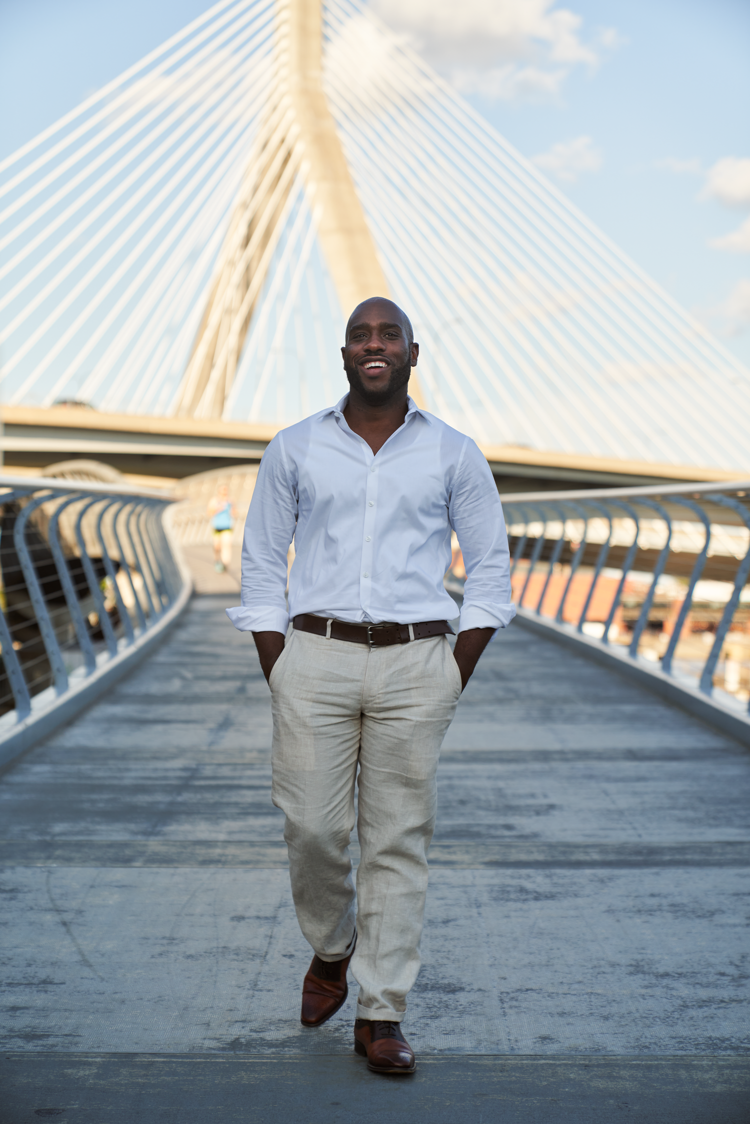 Smiling Black man in white shirt and khaki pants walking confidently on a bridge with cable-stayed structure behind him.