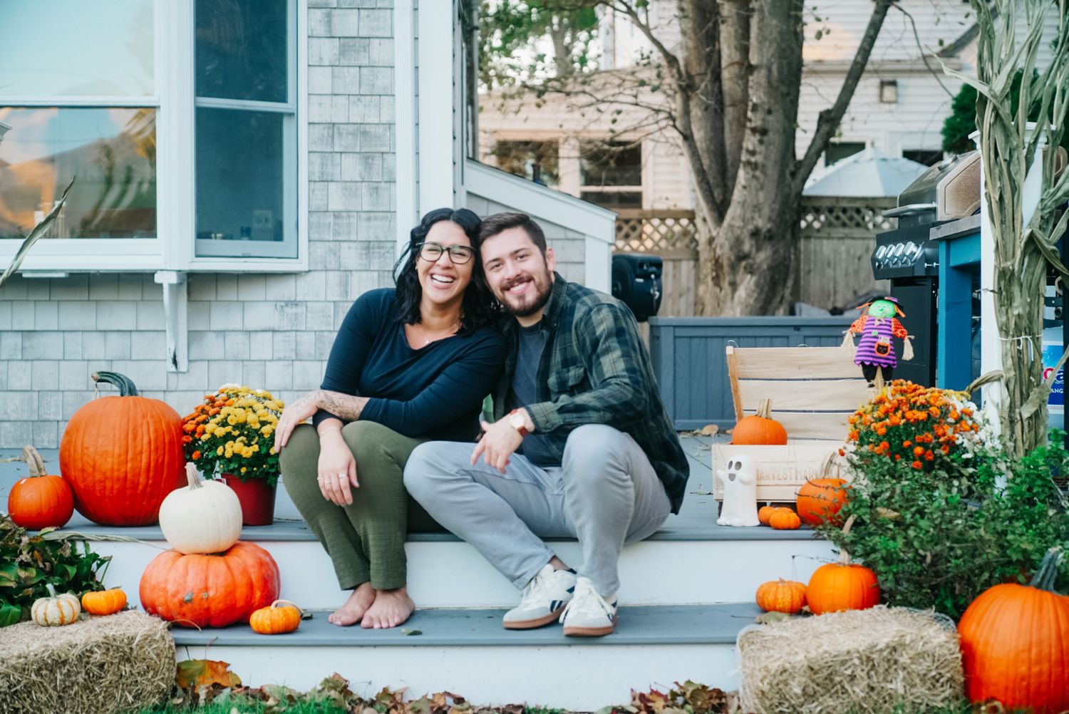 Happy couple posing together on home steps decorated with orange pumpkins and fall mums.