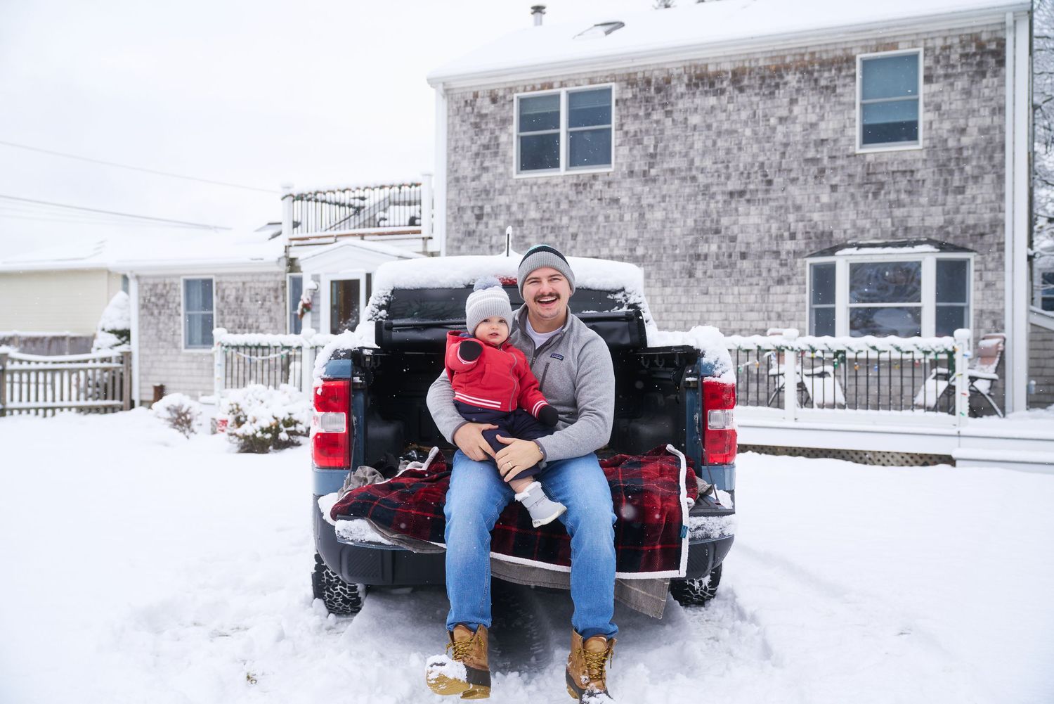Man and baby sitting on truck tailgate in snowy winter weather outside a house.