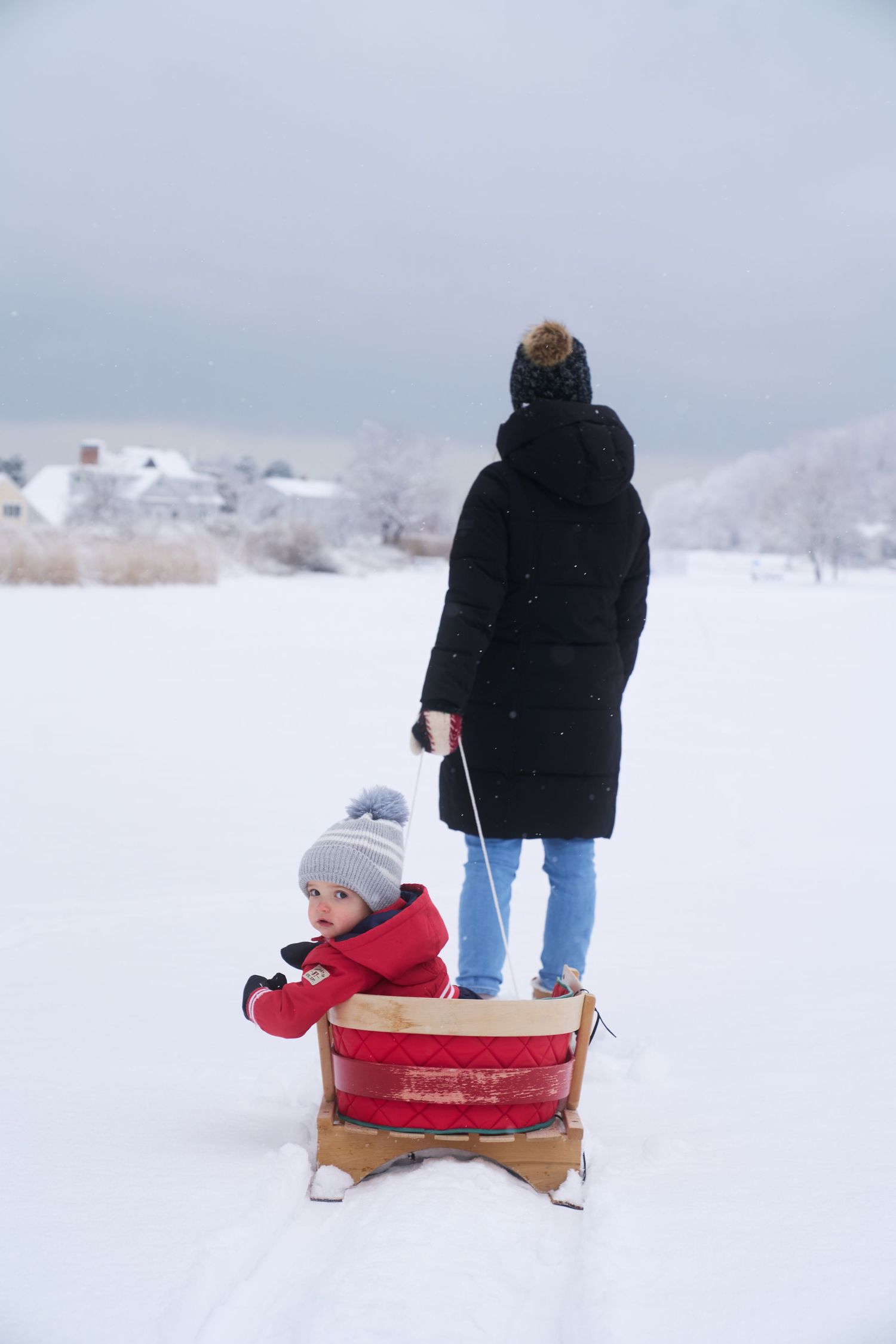 Mother in black coat pulling toddler in red snowsuit on a classic wooden red sled in snow.