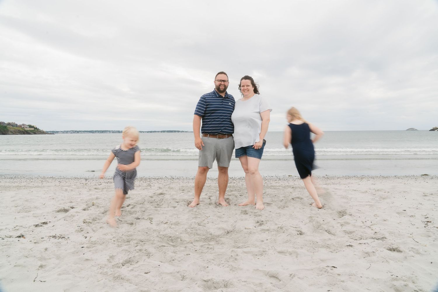 Family of four posing on a sandy beach with ocean waves in the background on a cloudy day.