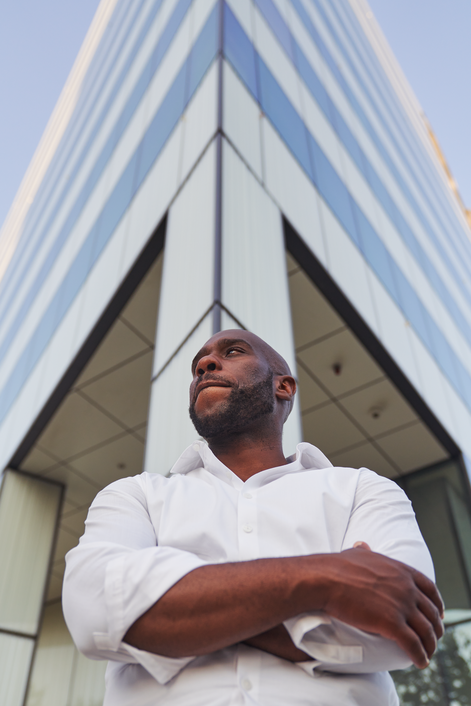 Confident Black businessman in white shirt standing arms crossed before modern glass office building, looking up.